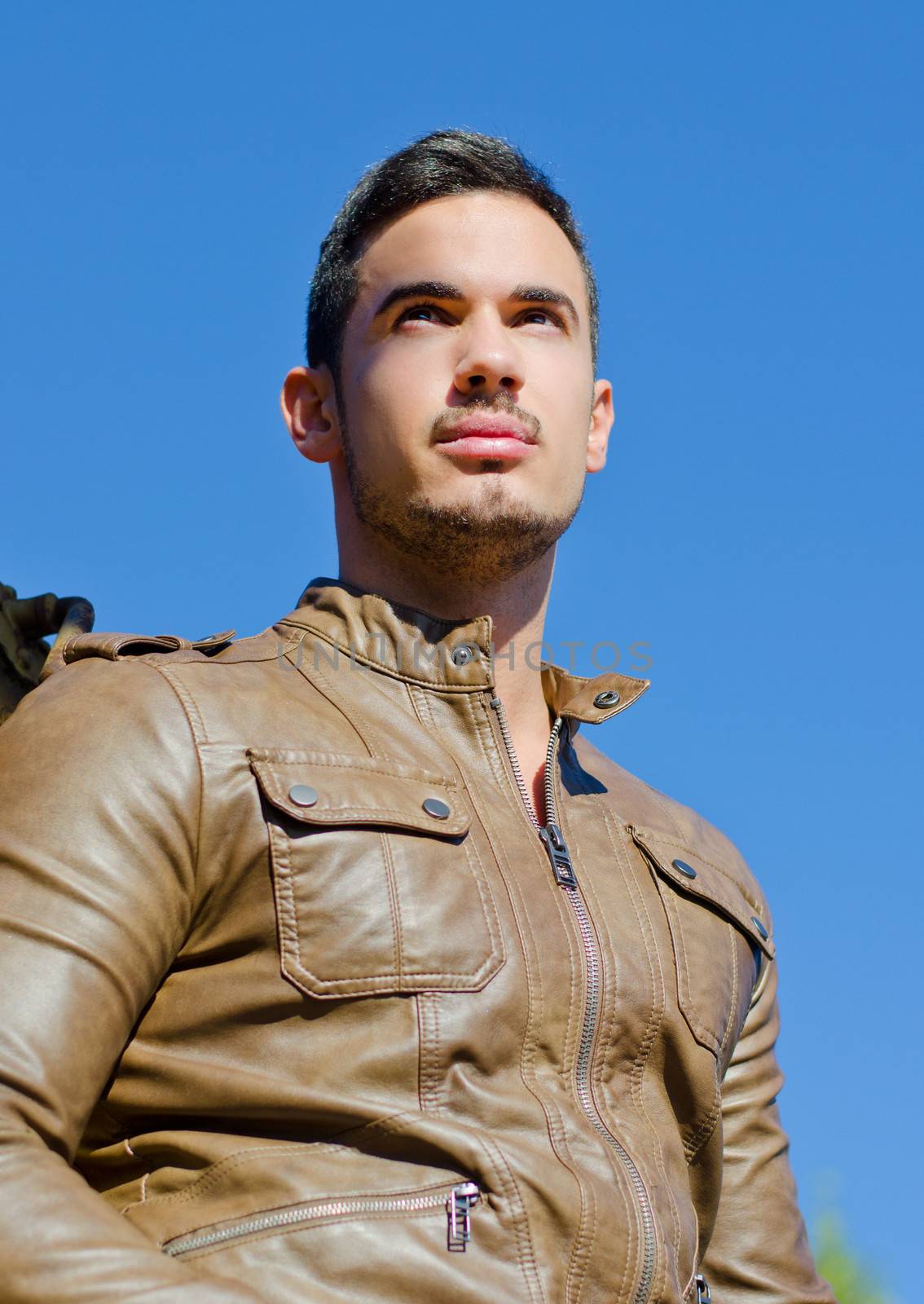 Handsome young man in leather jacket against blue sky looking far, shot from below