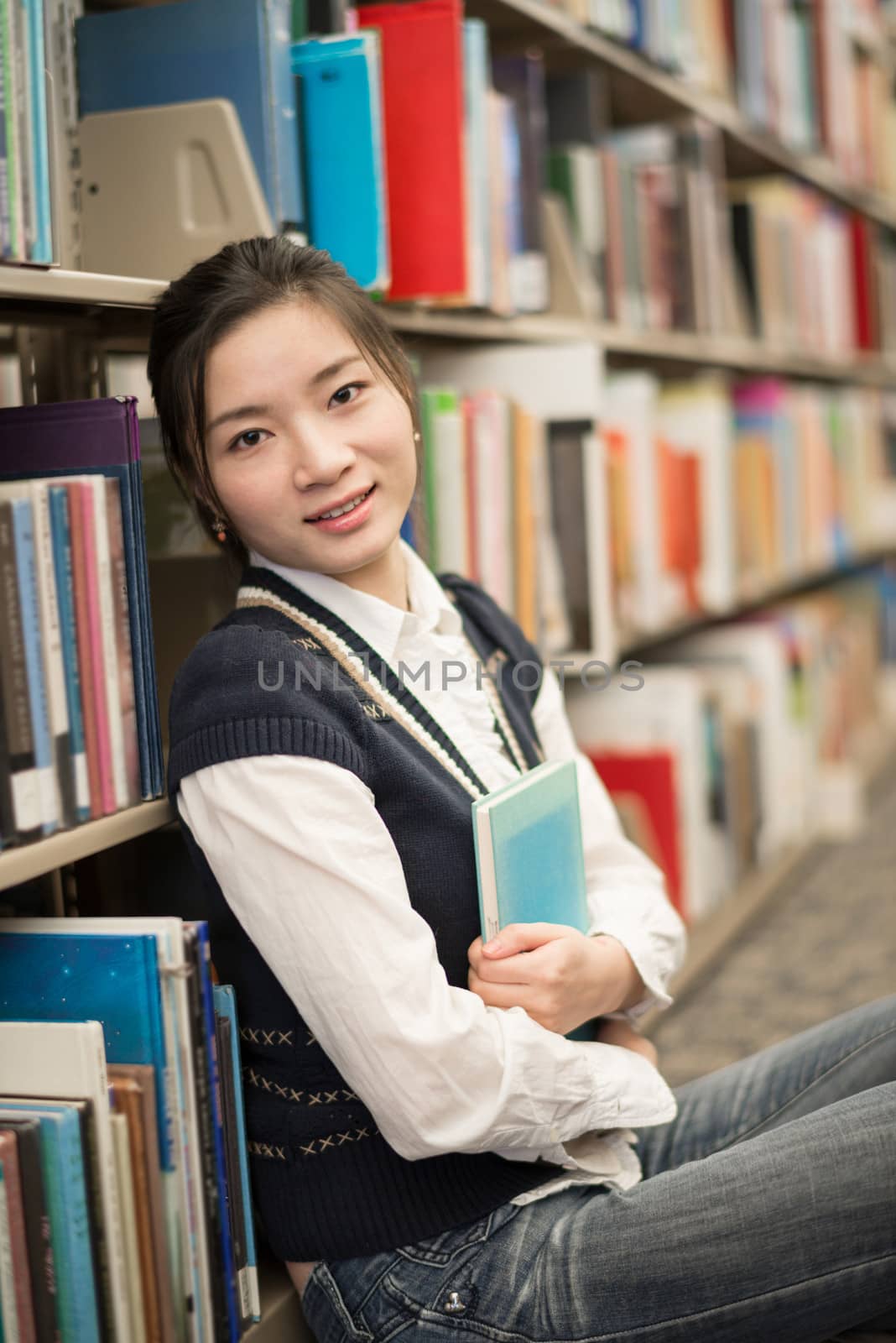 Cute young girl hugging a blue book while sitting in front of a bookshelf