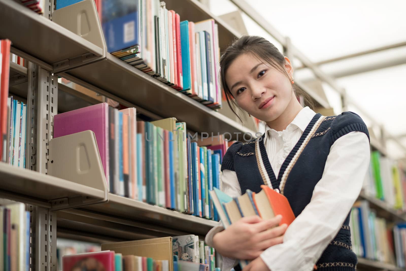 Portrait of cute young female student holding a stack of books next to a bookshelf in library