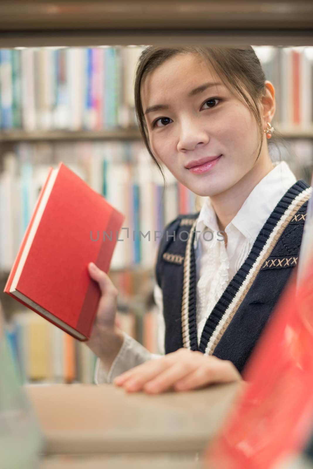 Pretty young girl picking out a red book from a pile of books on a bookshelf in library