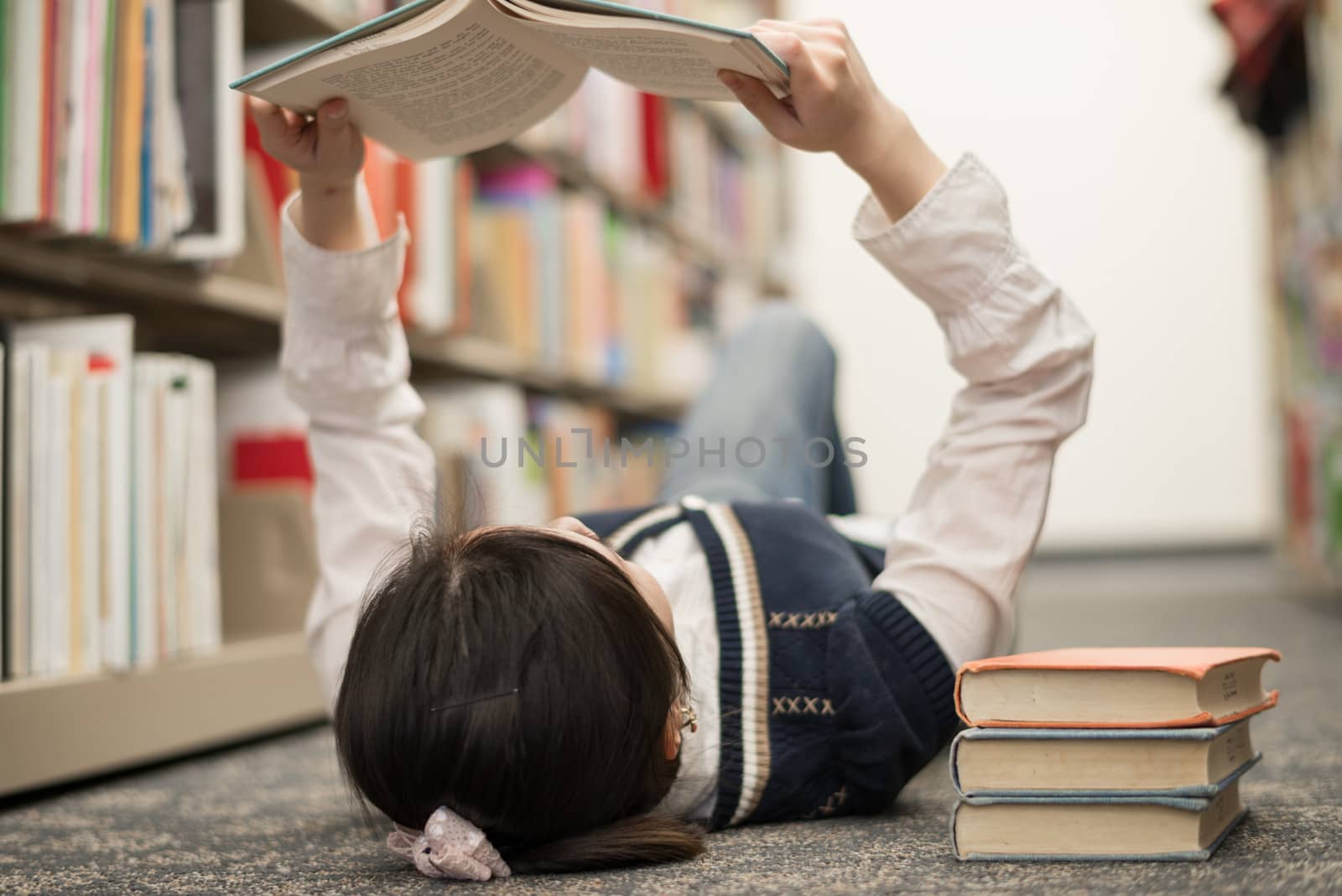 Young girl student laying on the library floor and reading a stack of books