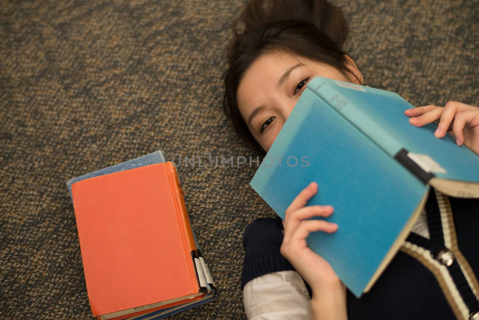 Fun young student laying on carpet with a book covering mouth next to a stack of books