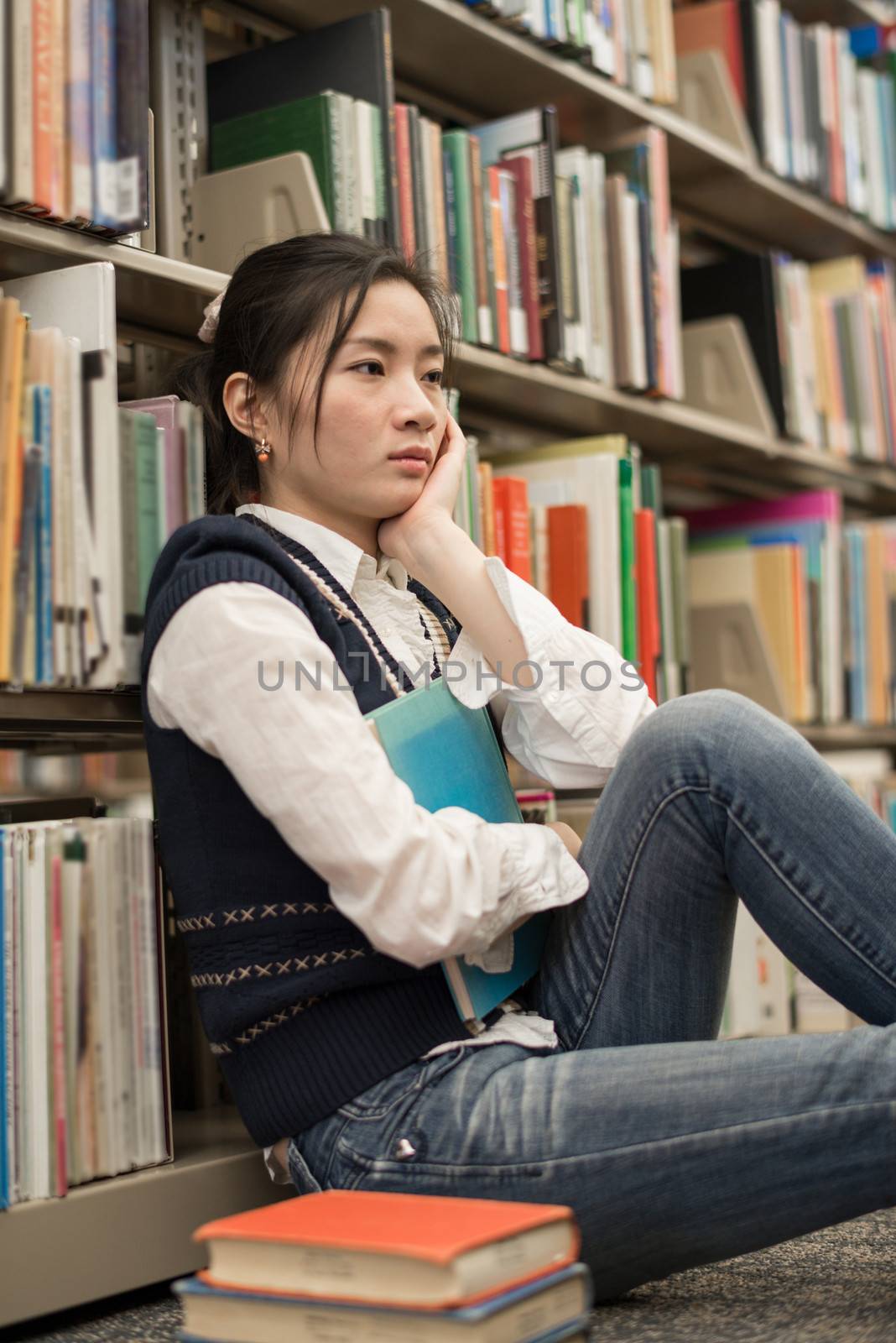 Young female student sitting near a stack of books and a bookshelf looking depressed