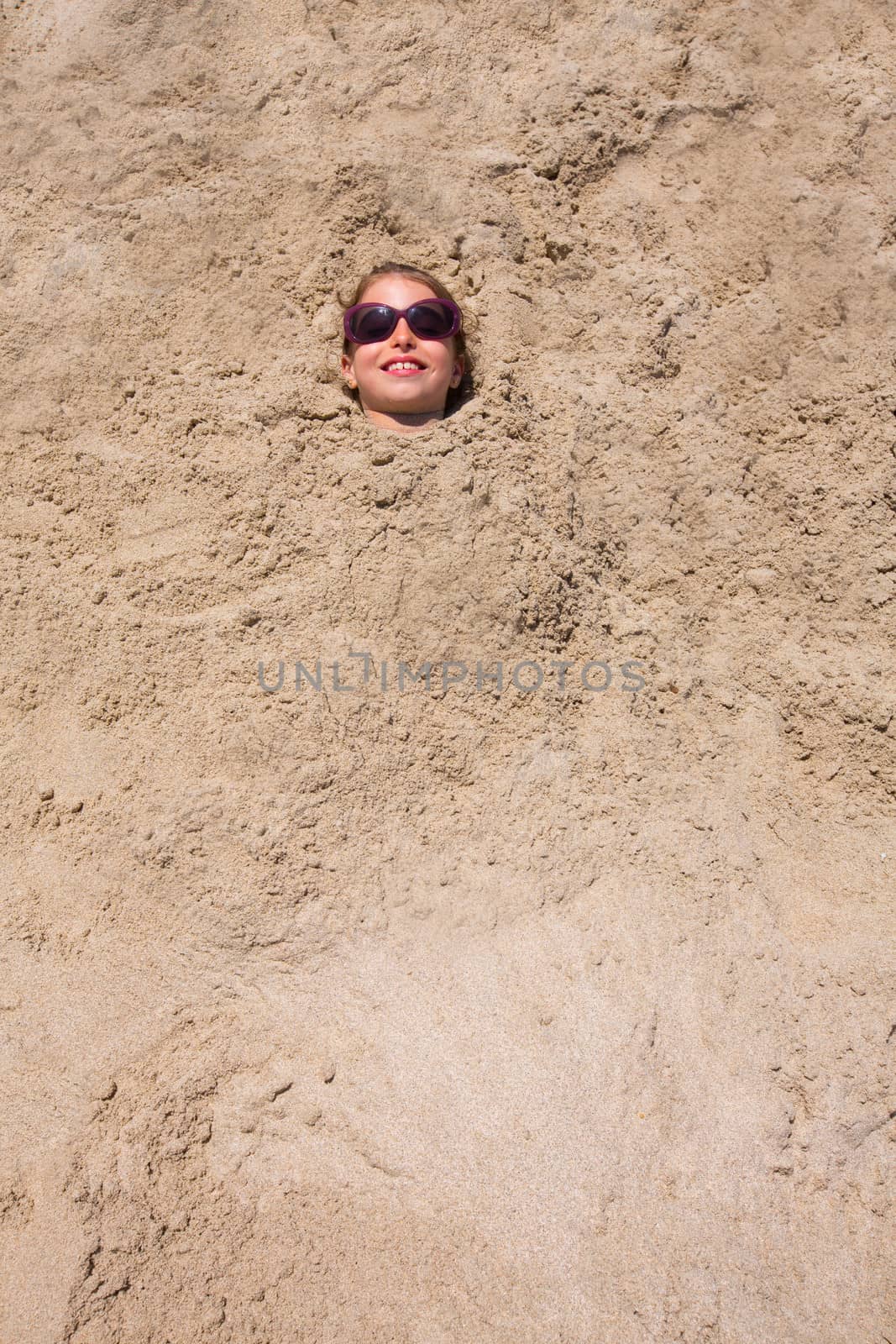 Funny kid girl playing buried in beach sand smiling with sunglasses at summer vacation