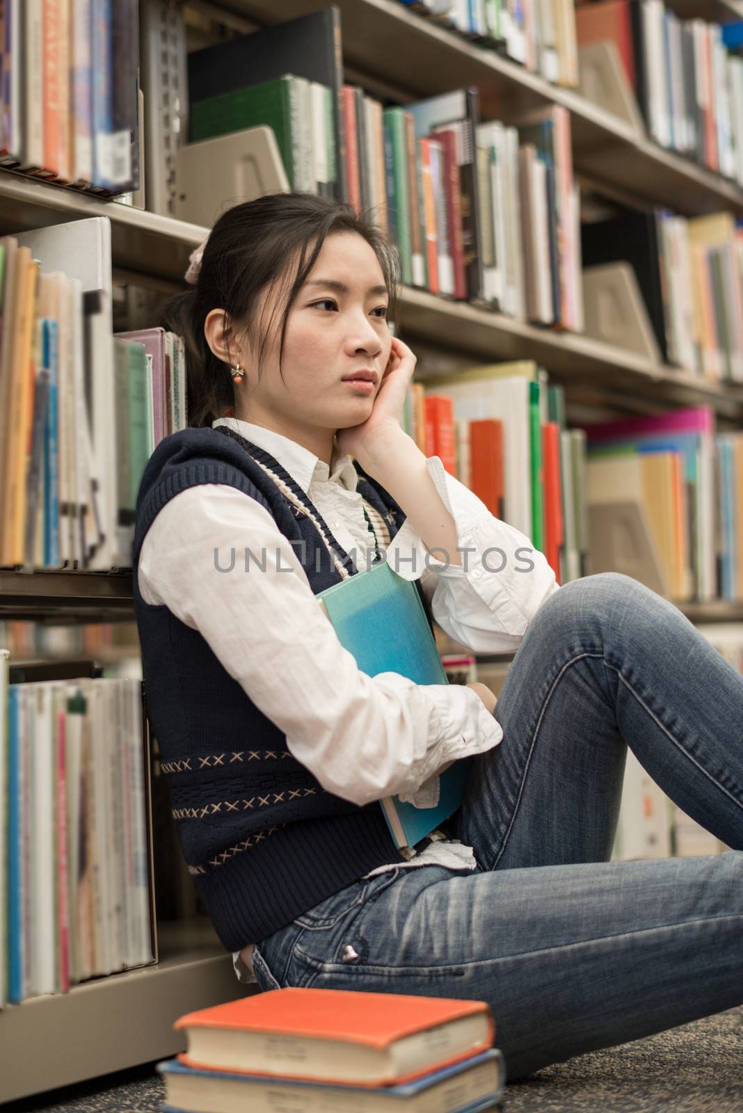 Young female student sitting near a stack of books and a bookshelf looking depressed