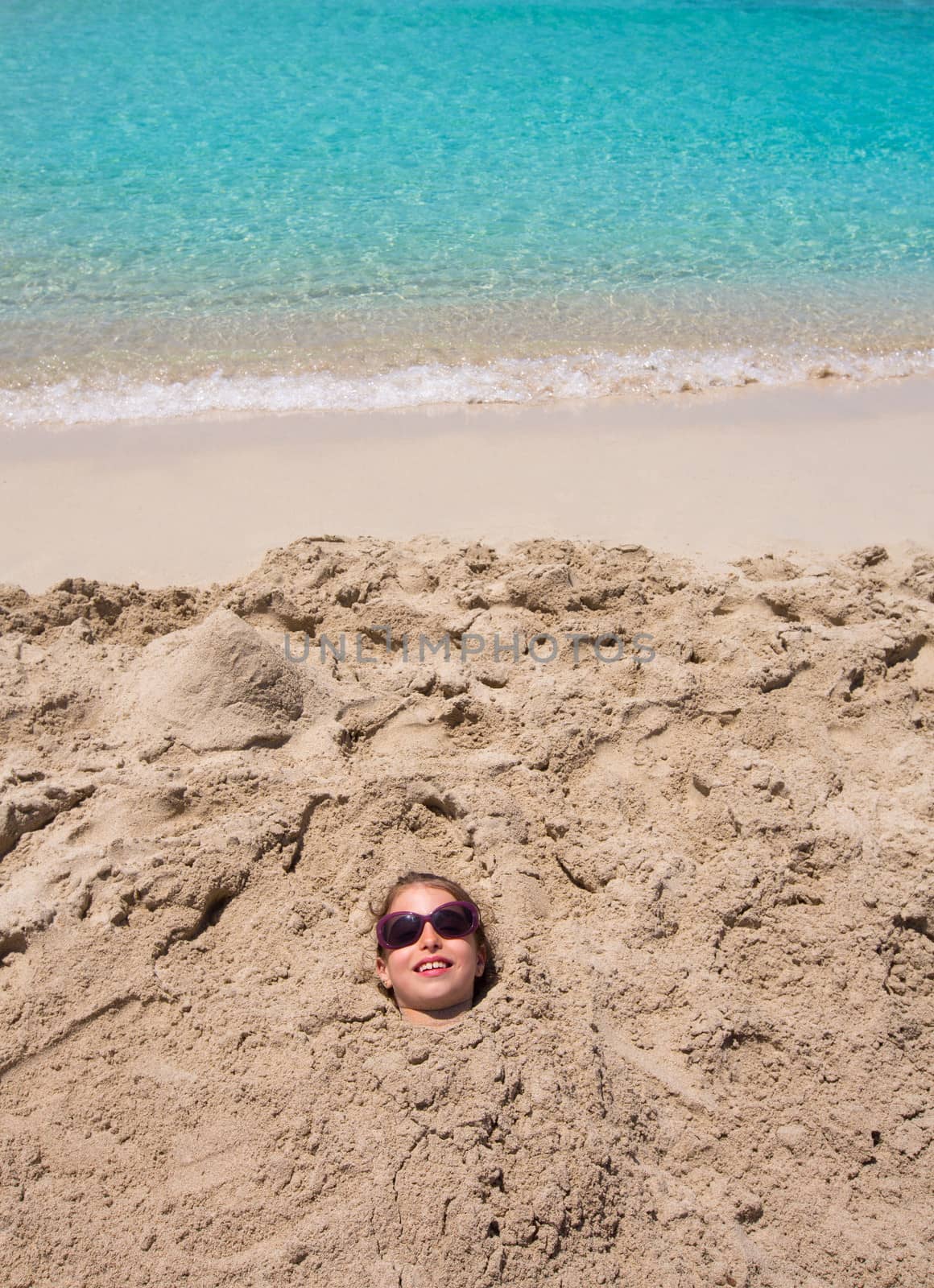 Funny kid girl playing buried in beach sand smiling with sunglasses at summer vacation