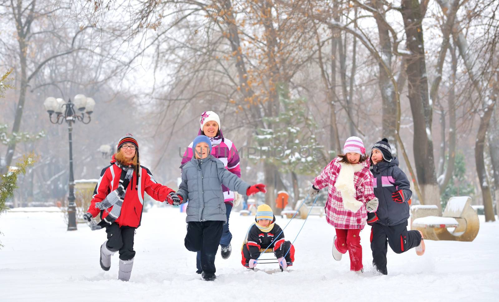 group of children and adult playing on snow in winter time, young girl pulling sister through snow on sled