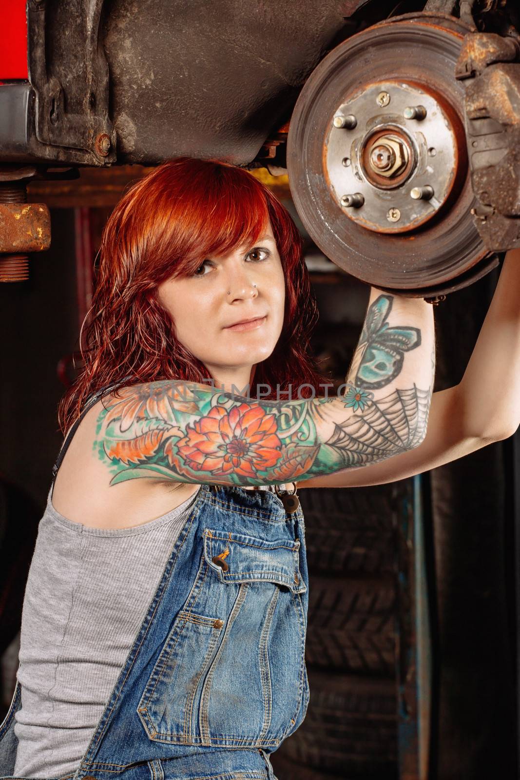 Photo of a young beautiful redhead mechanic wearing overalls and working on a car up on a hoist. Attached property release is for arm tattoos.

