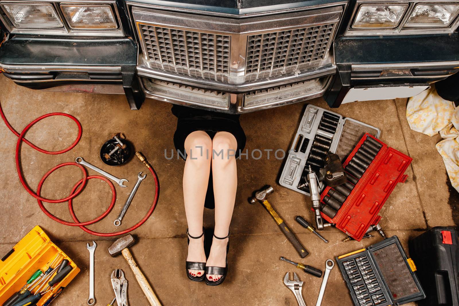 A woman wearing a black skirt and heels doing repairs under the front of an old car from the early 80's.