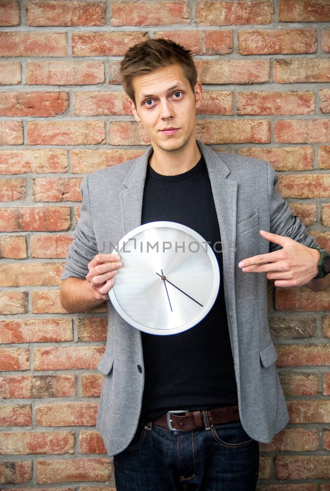 Young businessman holding a clock on brick wall background. Time in business concept