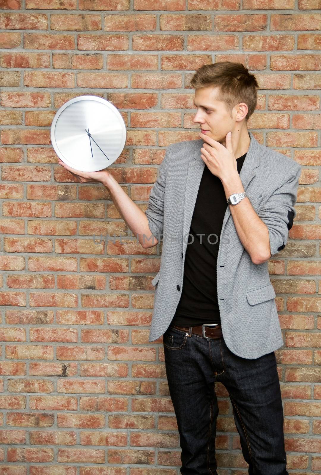 Young businessman holding a clock on brick wall background. Time in business concept