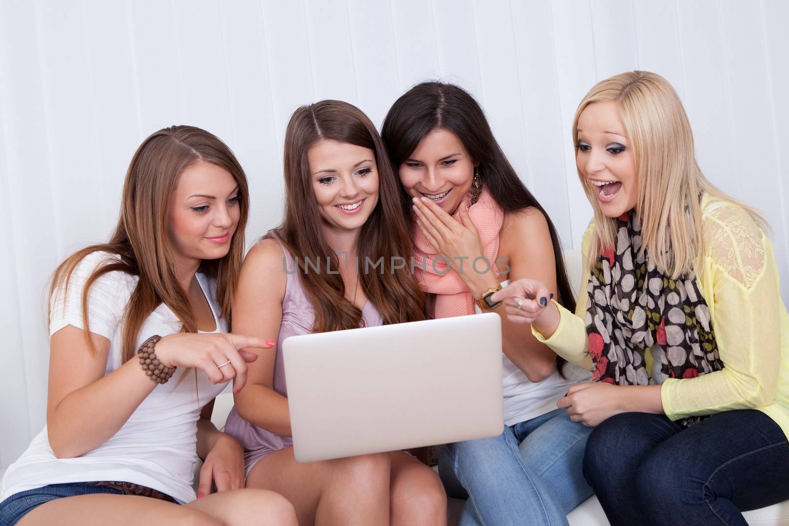 Group of beautiful young women sitting together on a couch sharing a laptop and smiling at something on the screen