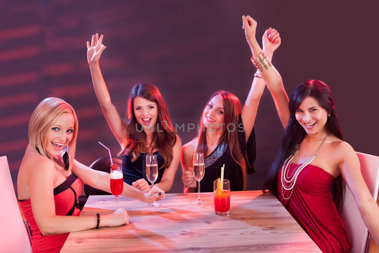 Glamorous young women celebrating in a nightclub sitting around a table laughing and raising their arms in the air in jubilation