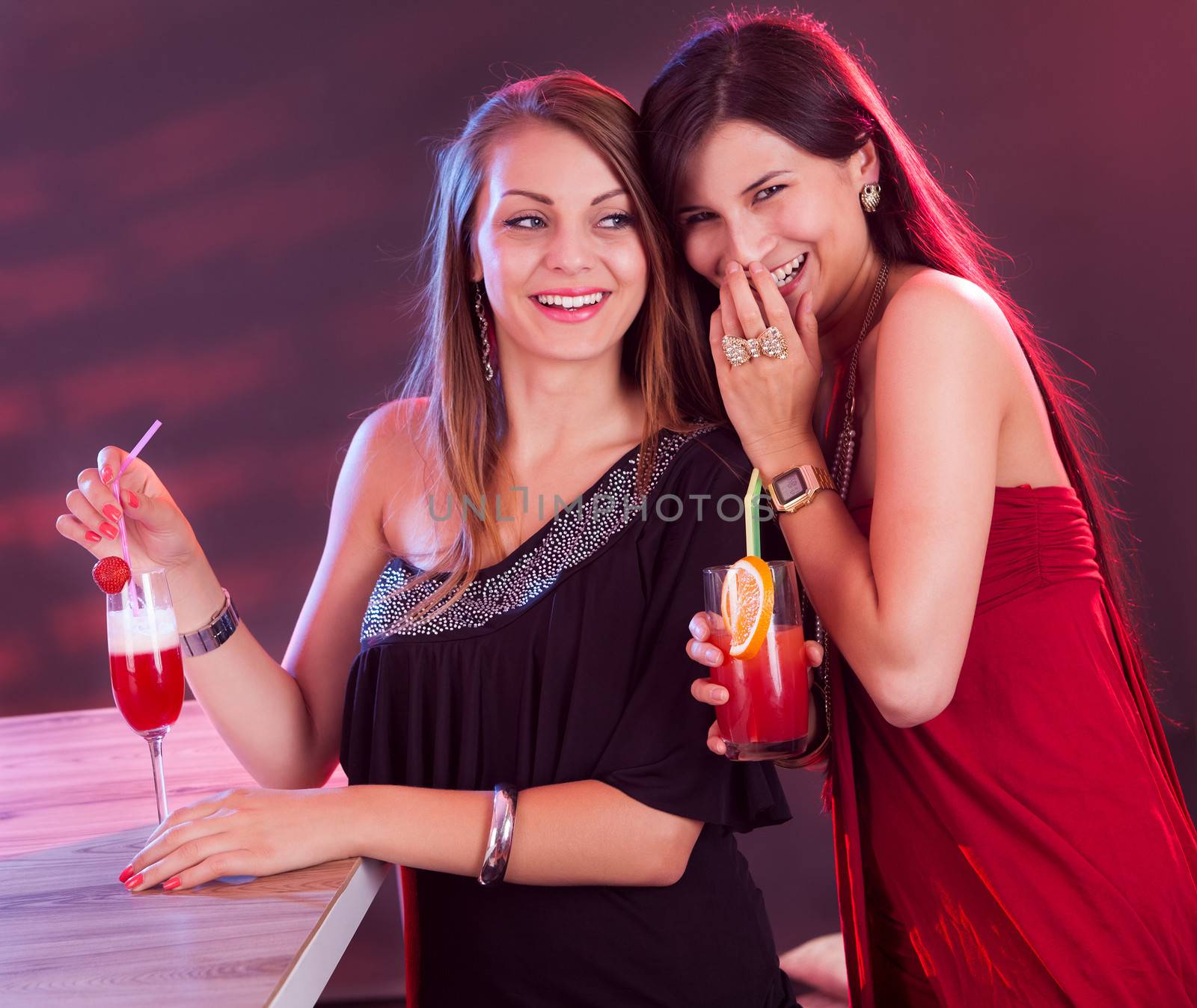 Two beautiful long haired female friends partying at a bar counter with cocktails in their hands under colourful lightiing in a nightclub