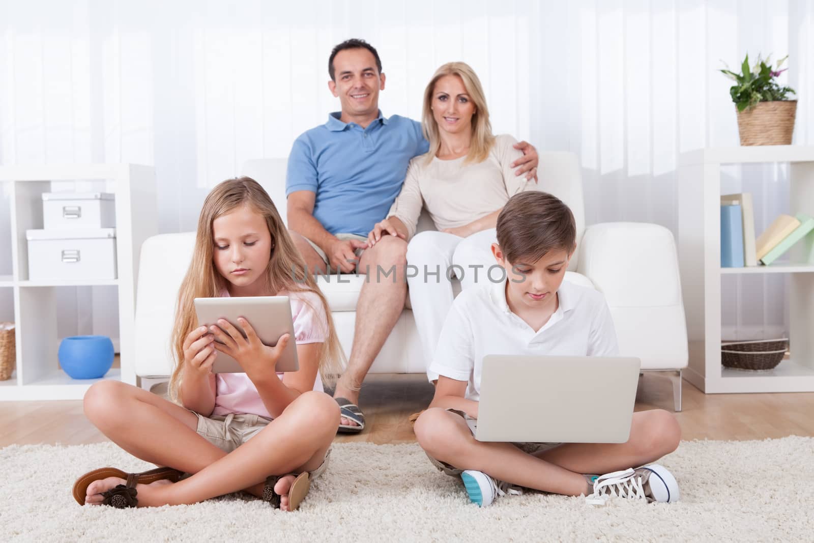 Children On The Carpet Using Tablet And Laptop With Parents Behind Them At Home