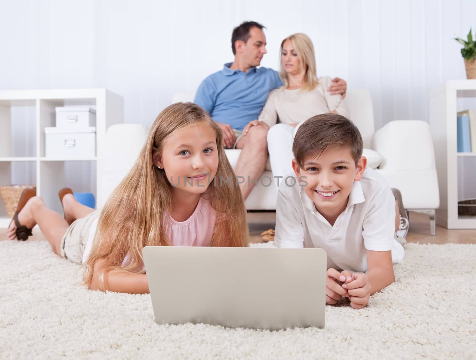 Children On The Carpet Using Tablet And Laptop With Parents Behind Them At Home