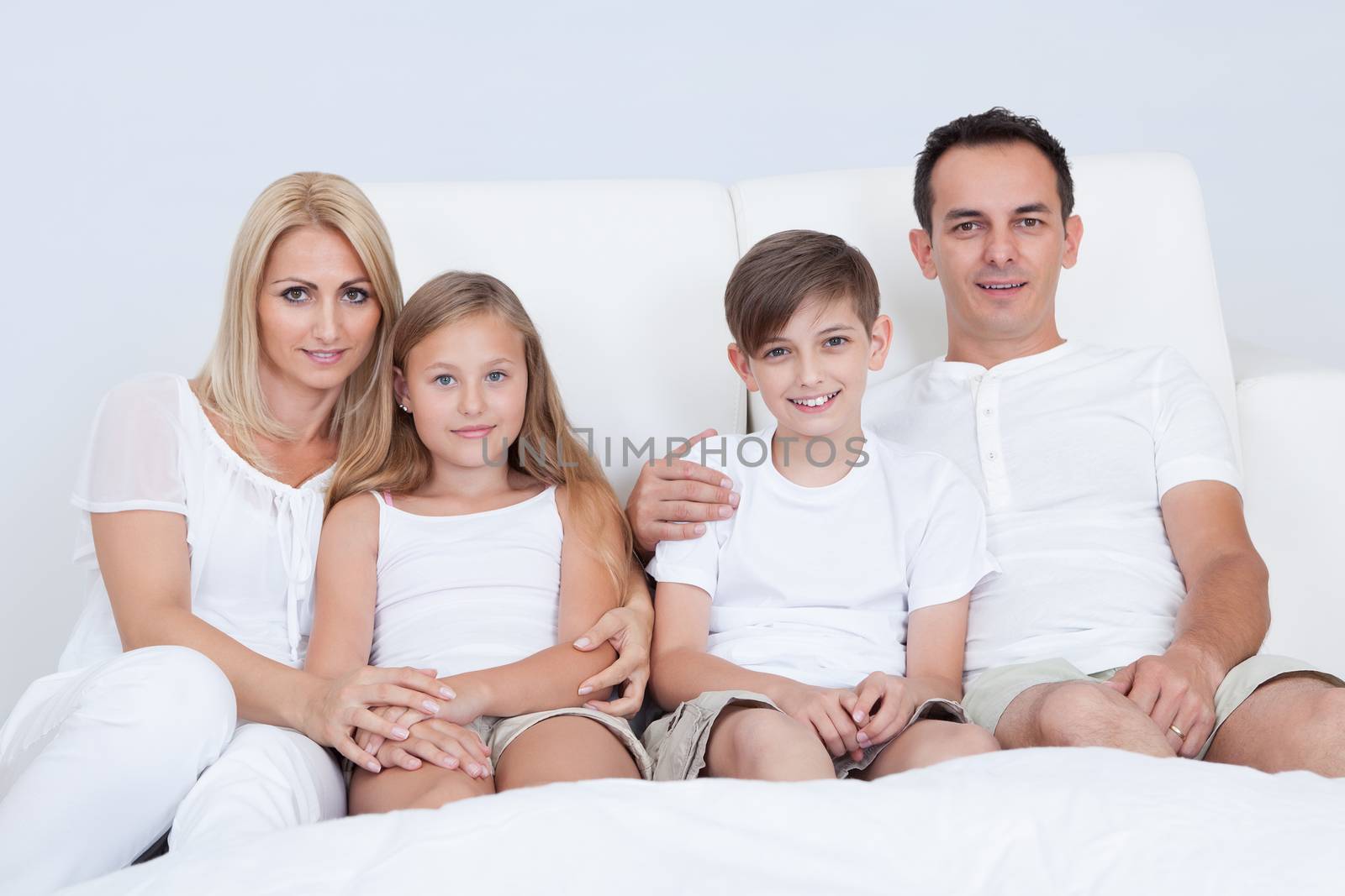 Portrait Of Happy Family With Two Children Sitting On Bed In Bedroom
