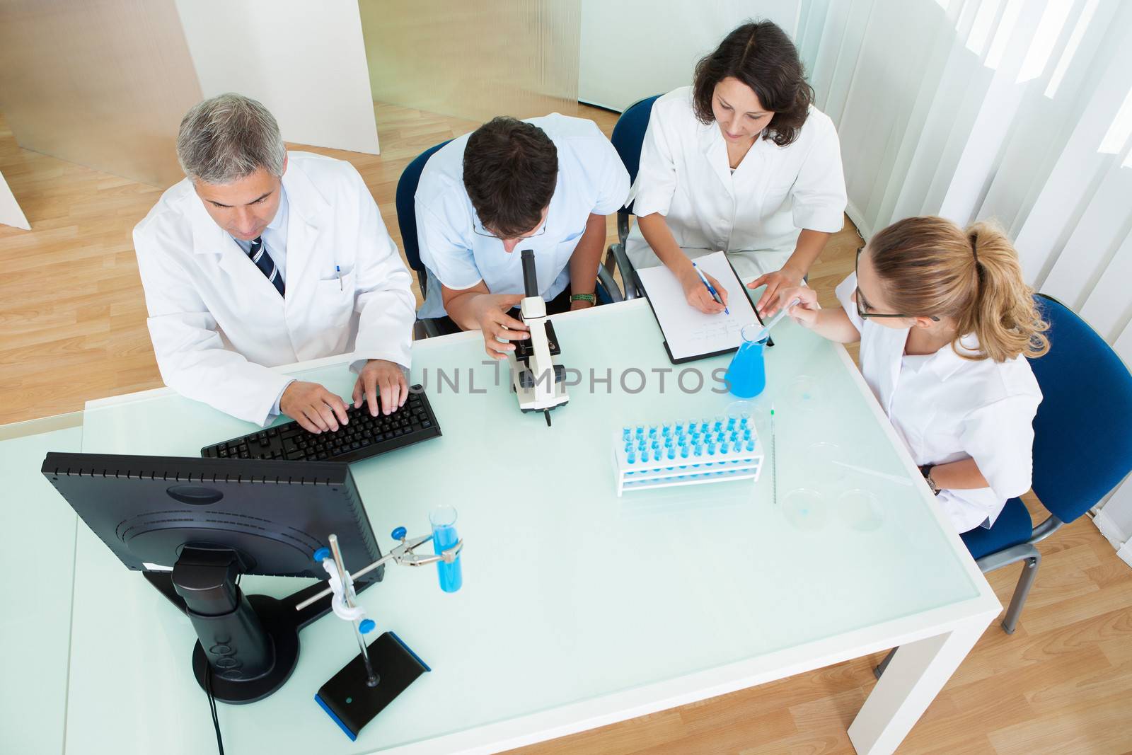 Overhead view of four laboratory technicians at work seated around a table reading and recording tests on a microscope and computer