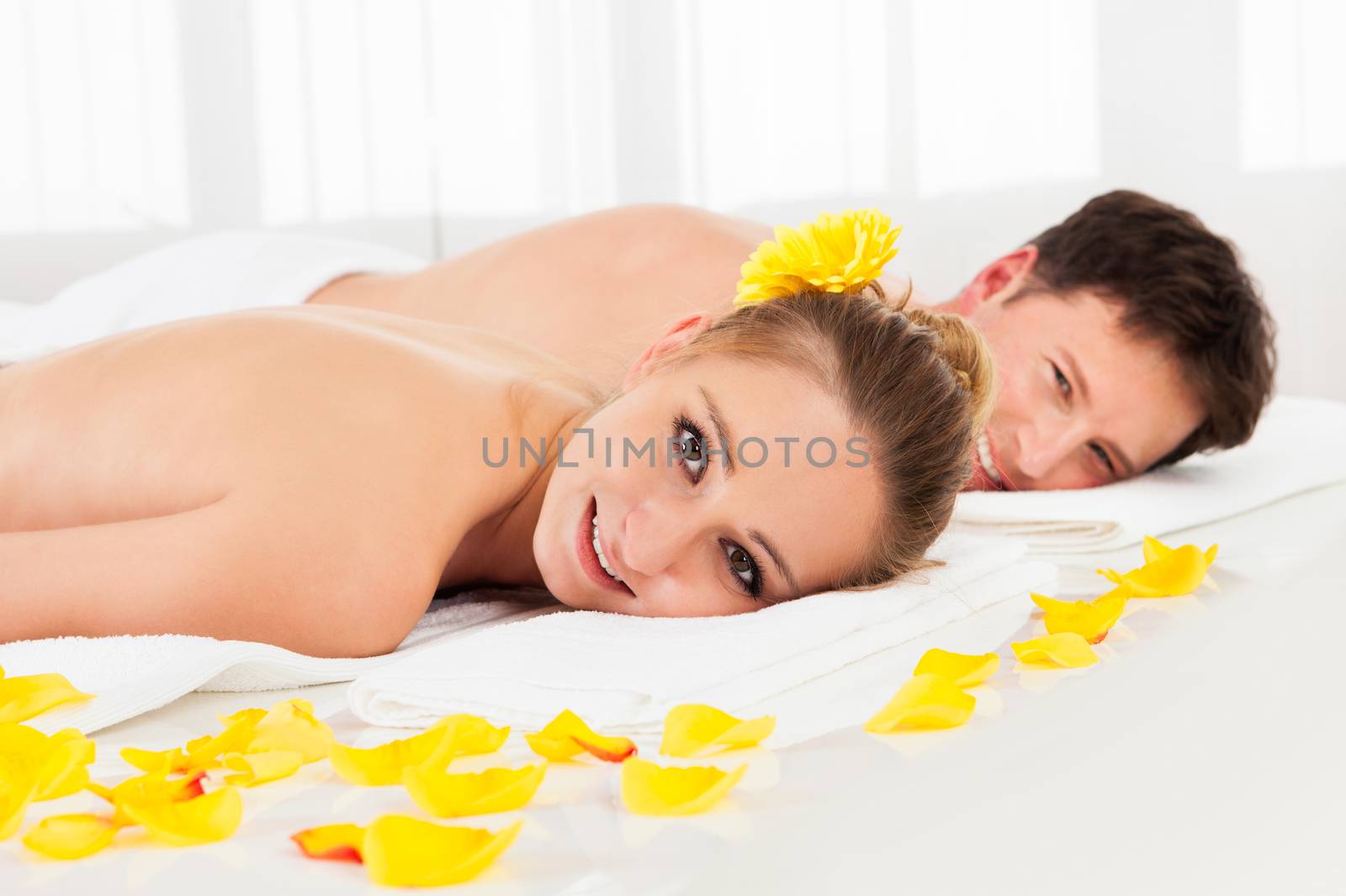 Smiling relaxed couple lying side by side on a table amongst yellow flowers at a spa with their top half bare and a towel around their loins