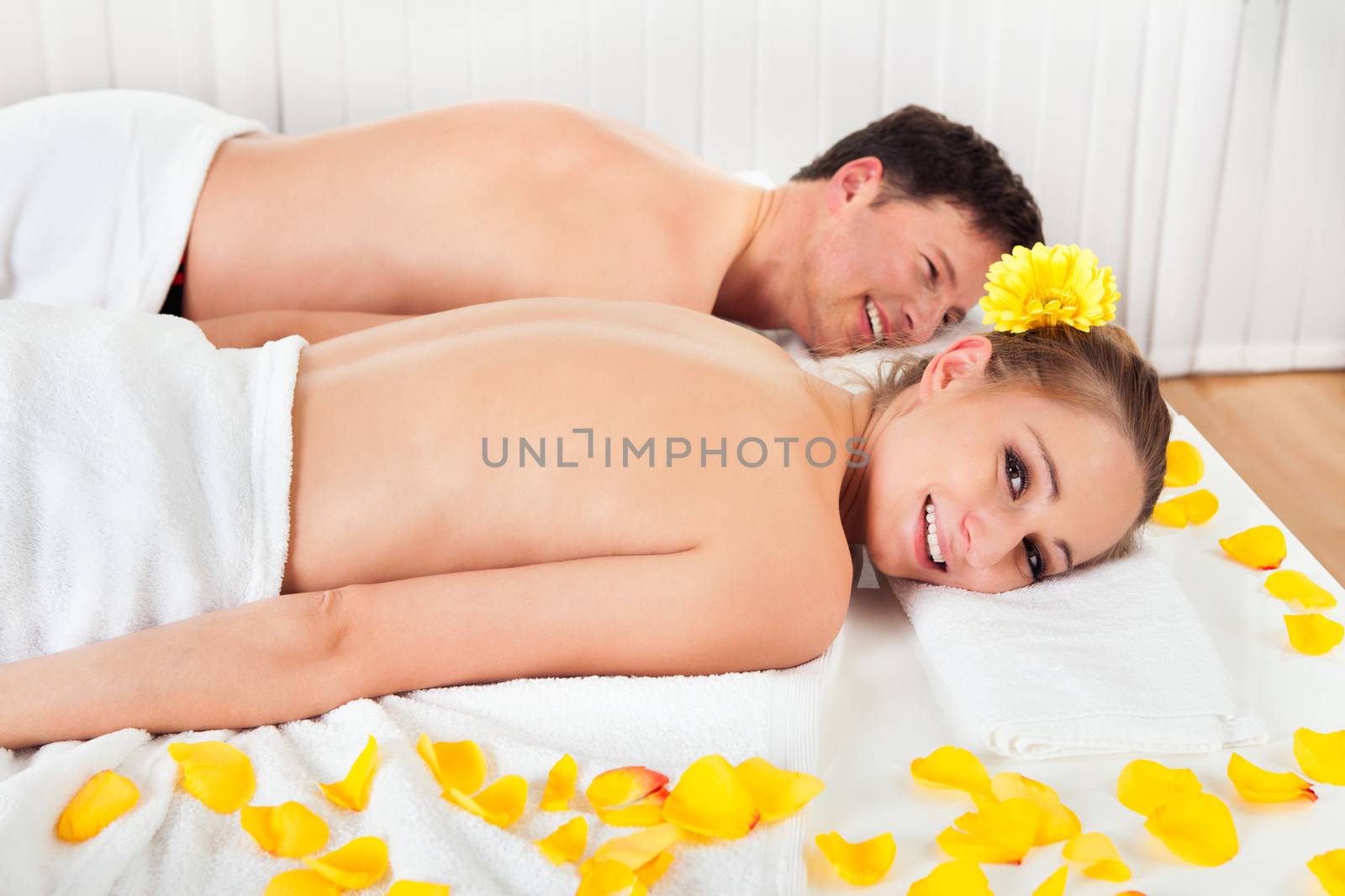 Smiling relaxed couple lying side by side on a table amongst yellow flowers at a spa with their top half bare and a towel around their loins