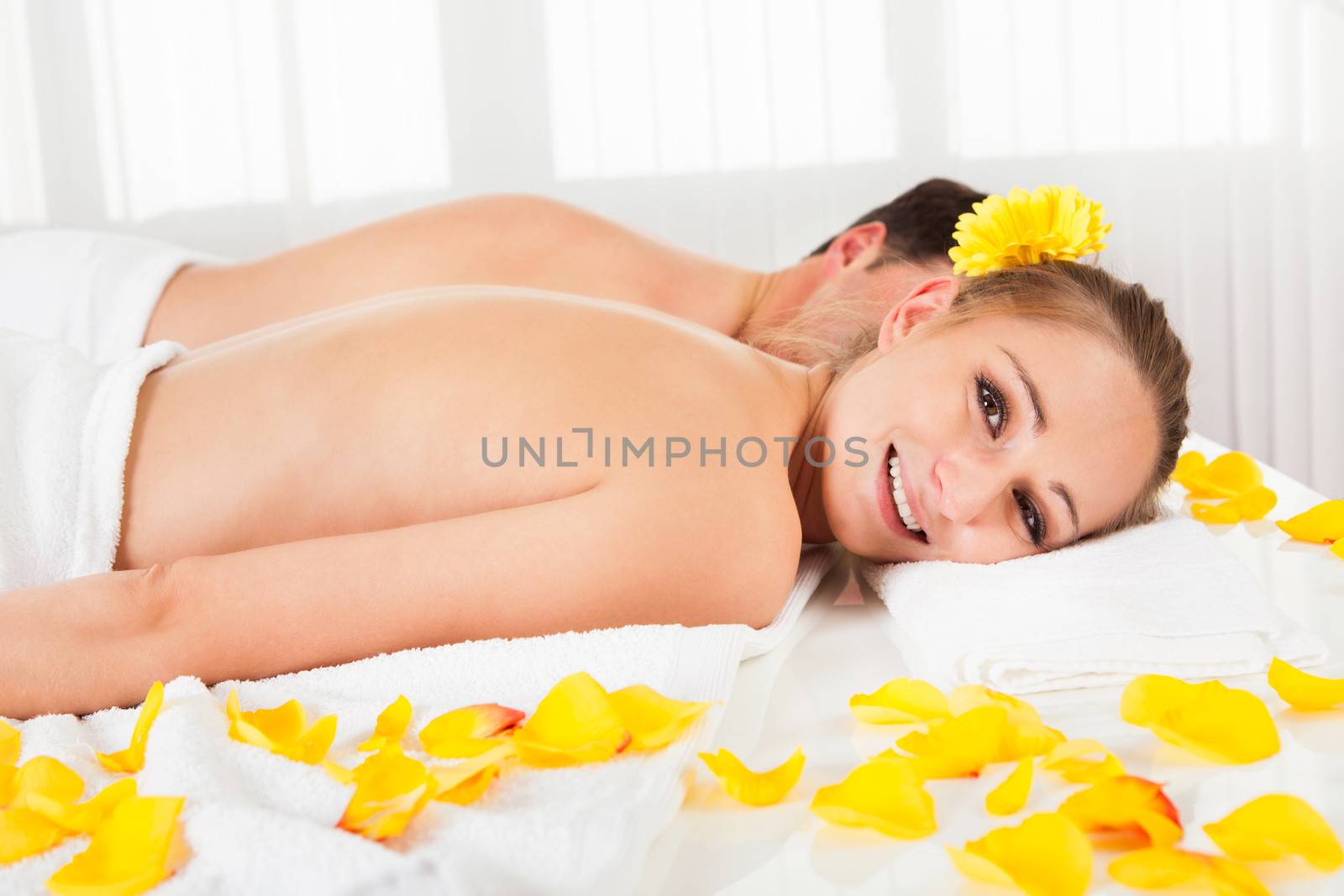 Smiling relaxed couple lying side by side on a table amongst yellow flowers at a spa with their top half bare and a towel around their loins