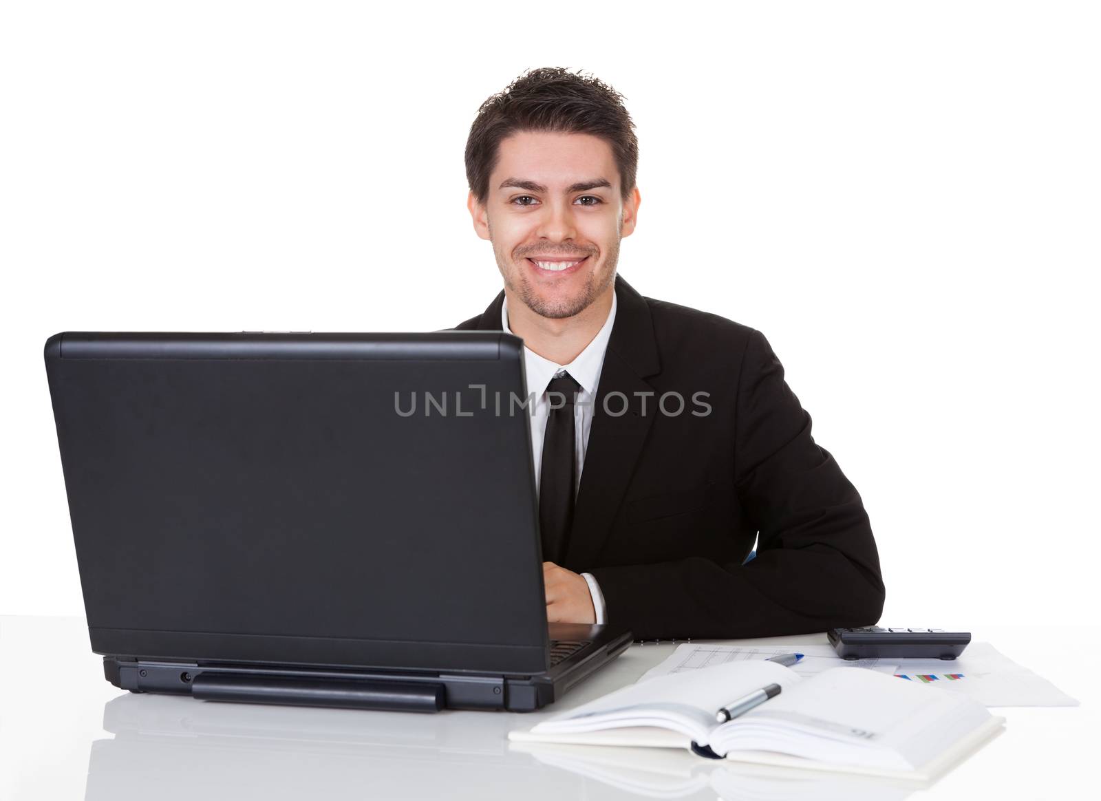 Smiling handsome young businessman seated at his desk behind his laptop computer looking at the camera