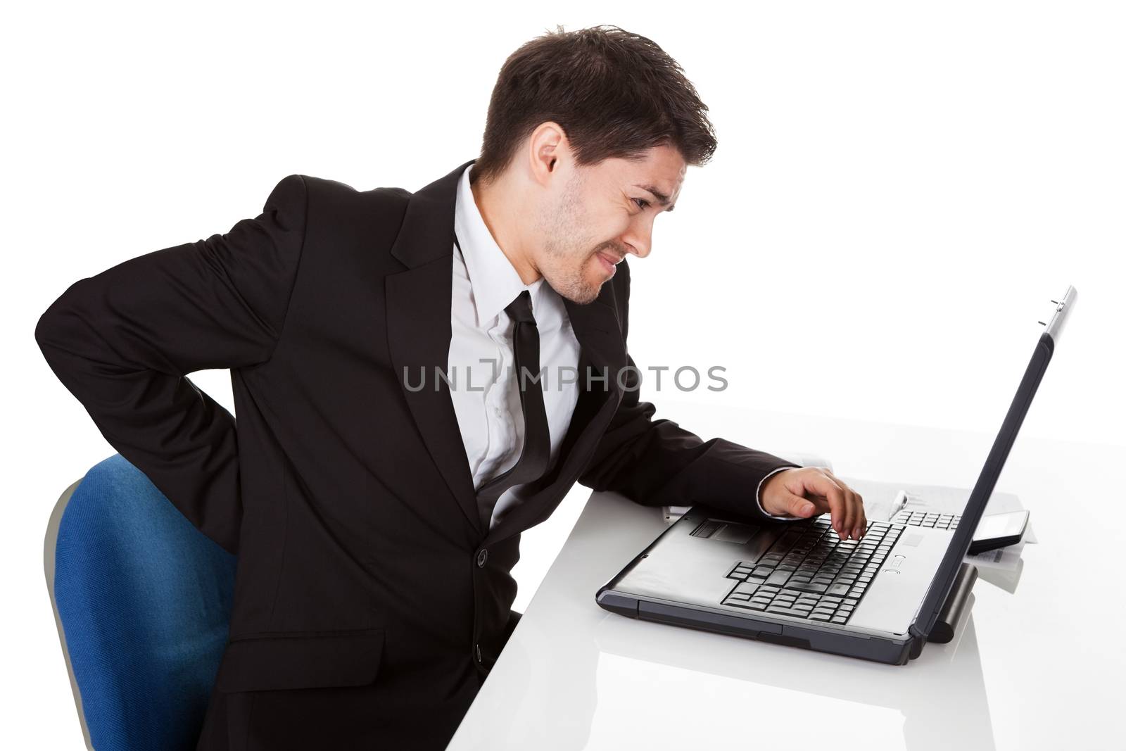 Businessman with lower back ache from sitting with a bad posture in his office chair working on his laptop massaging his back with his hand