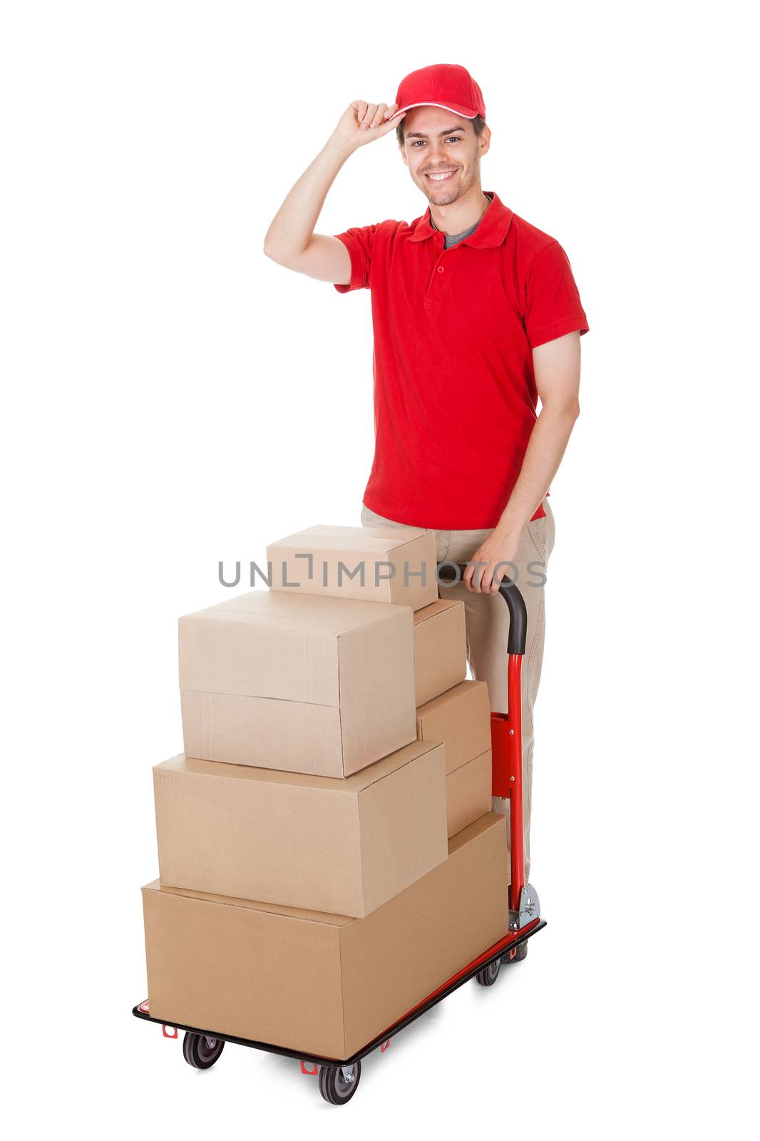 Cheerful young deliveryman in a red uniform holding trolley loaded with cardboard boxes isolated on white