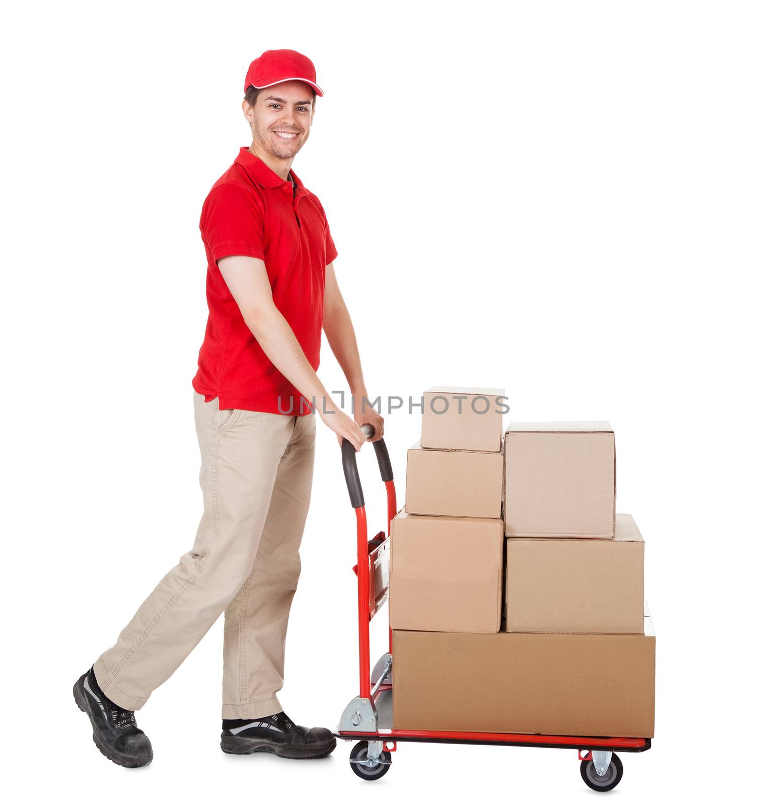 Cheerful young deliveryman in a red uniform holding trolley loaded with cardboard boxes isolated on white