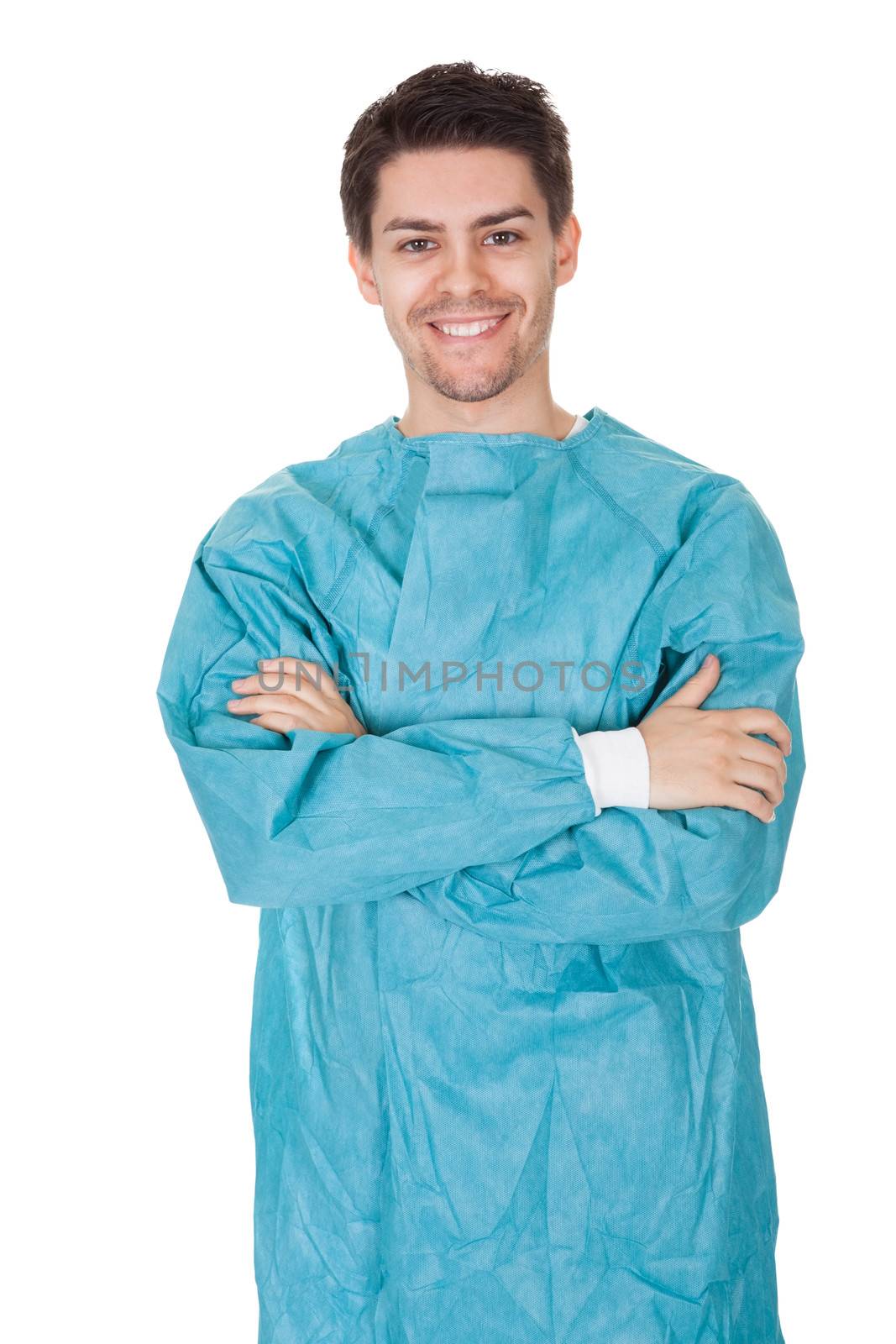 Smiling confident young surgeon wearing a gown and stethoscope standing with his arms crossed isolated on white