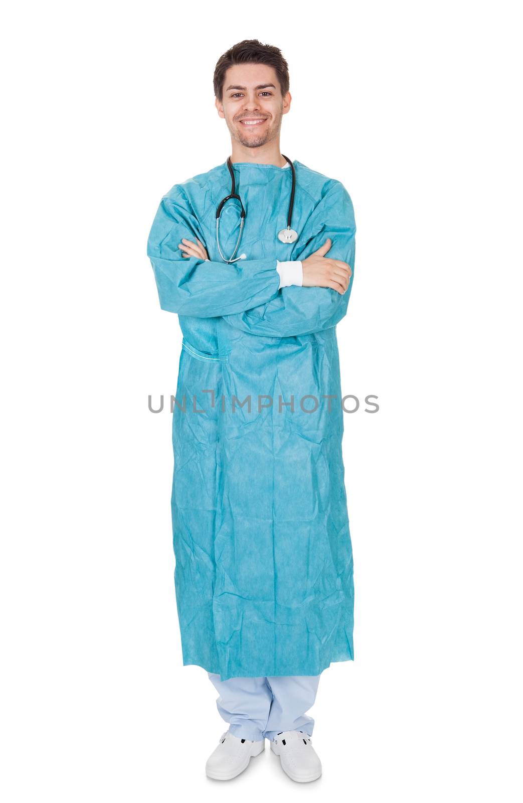 Smiling confident young surgeon wearing a gown and stethoscope standing with his arms crossed isolated on white