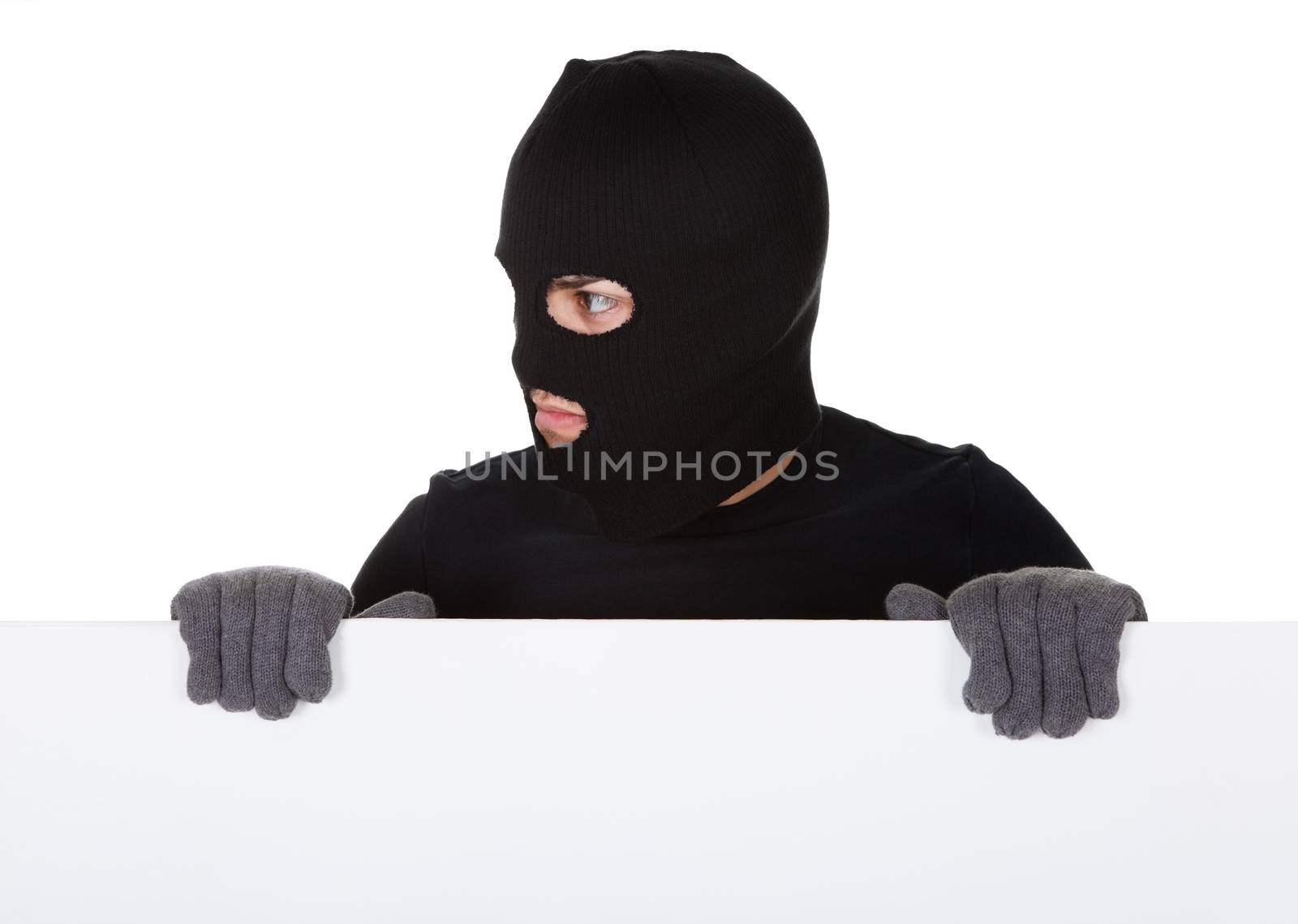 Thief in a balaclava and gloves looking around the edge of a blank sign with copyspace for your text isolated on white