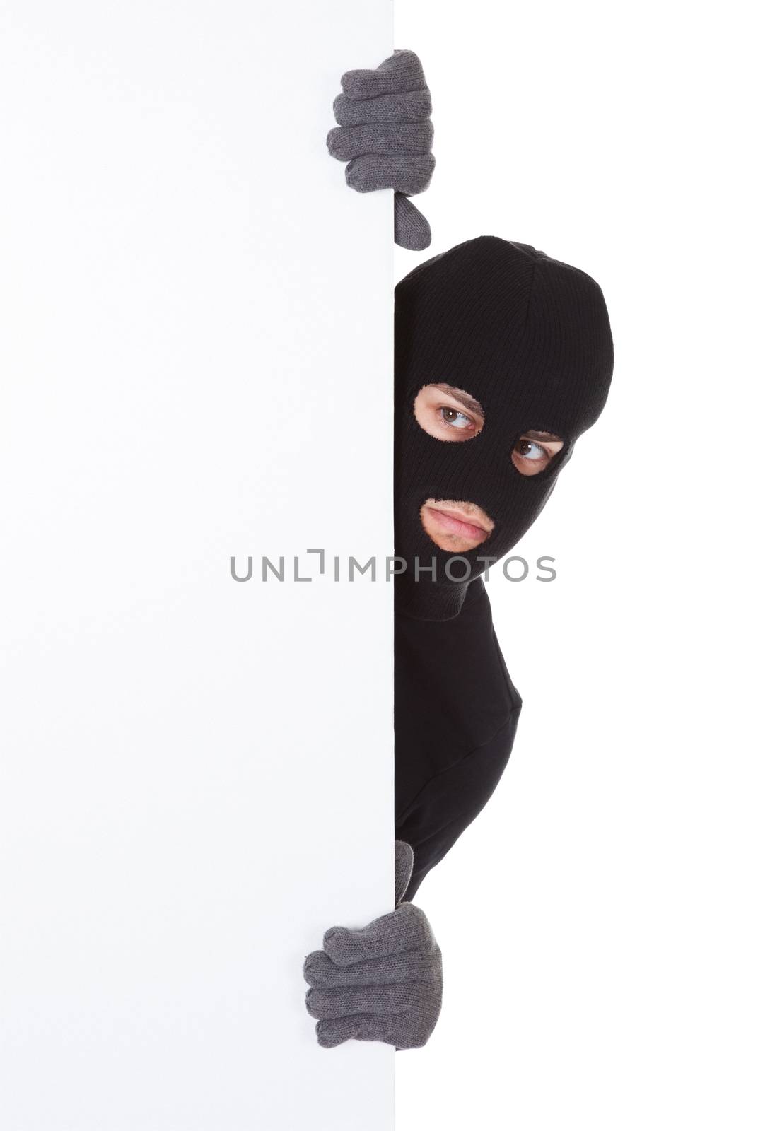 Thief in a balaclava and gloves looking around the edge of a blank sign with copyspace for your text isolated on white