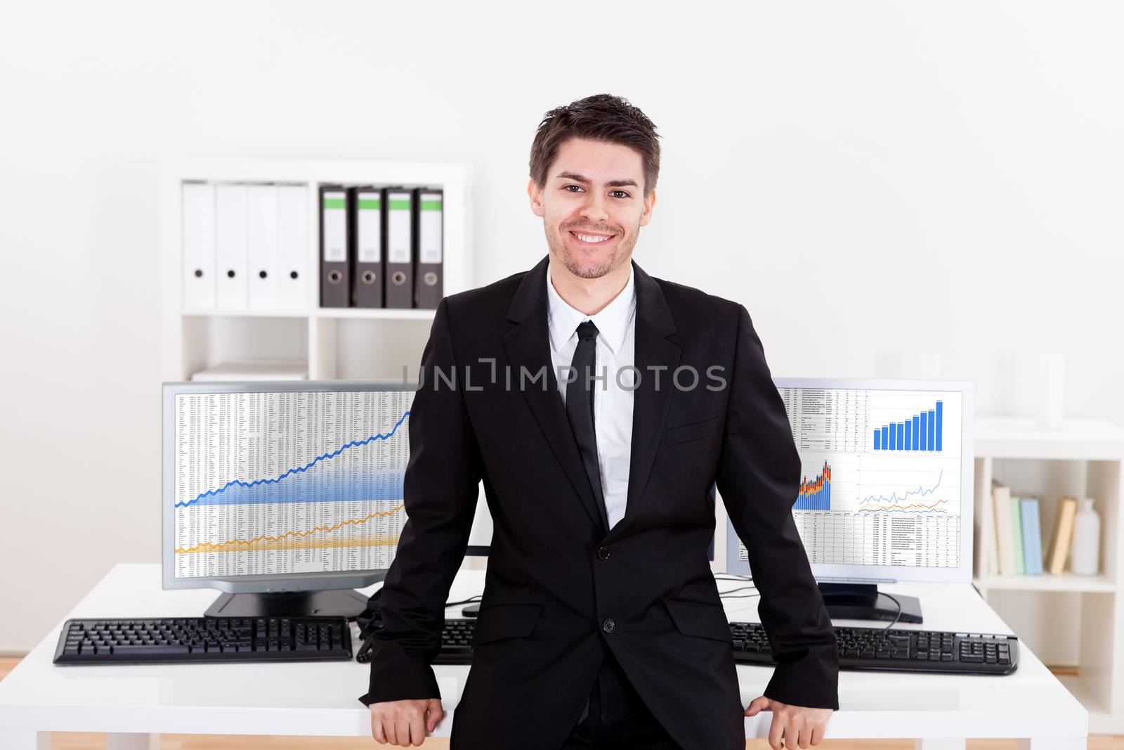 Confident smiling stock broker sitting on the edge of his desk surrounded by graphs and analytics indicating a successful bull market