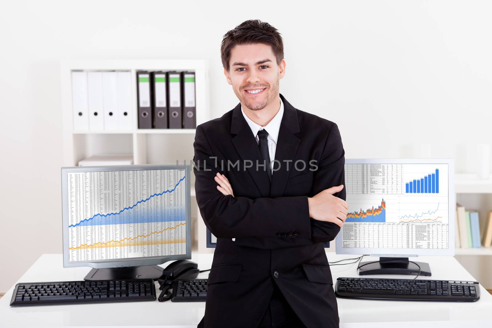 Confident smiling stock broker sitting on the edge of his desk surrounded by graphs and analytics indicating a successful bull market