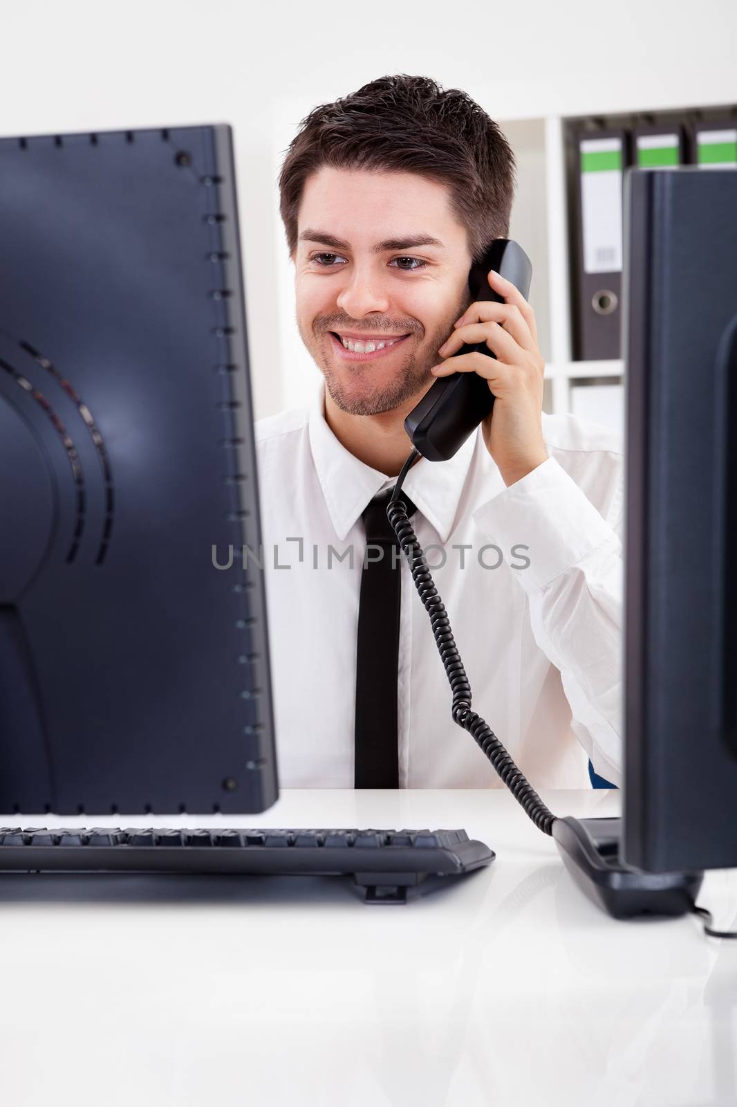 View between two computer monitors of a smiling handsome young stock broker talking on a phone