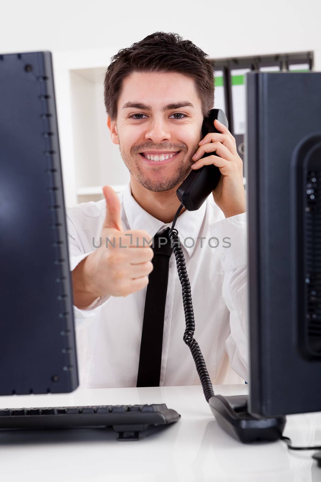 View between two computer monitors of a smiling handsome young stock broker talking on a phone