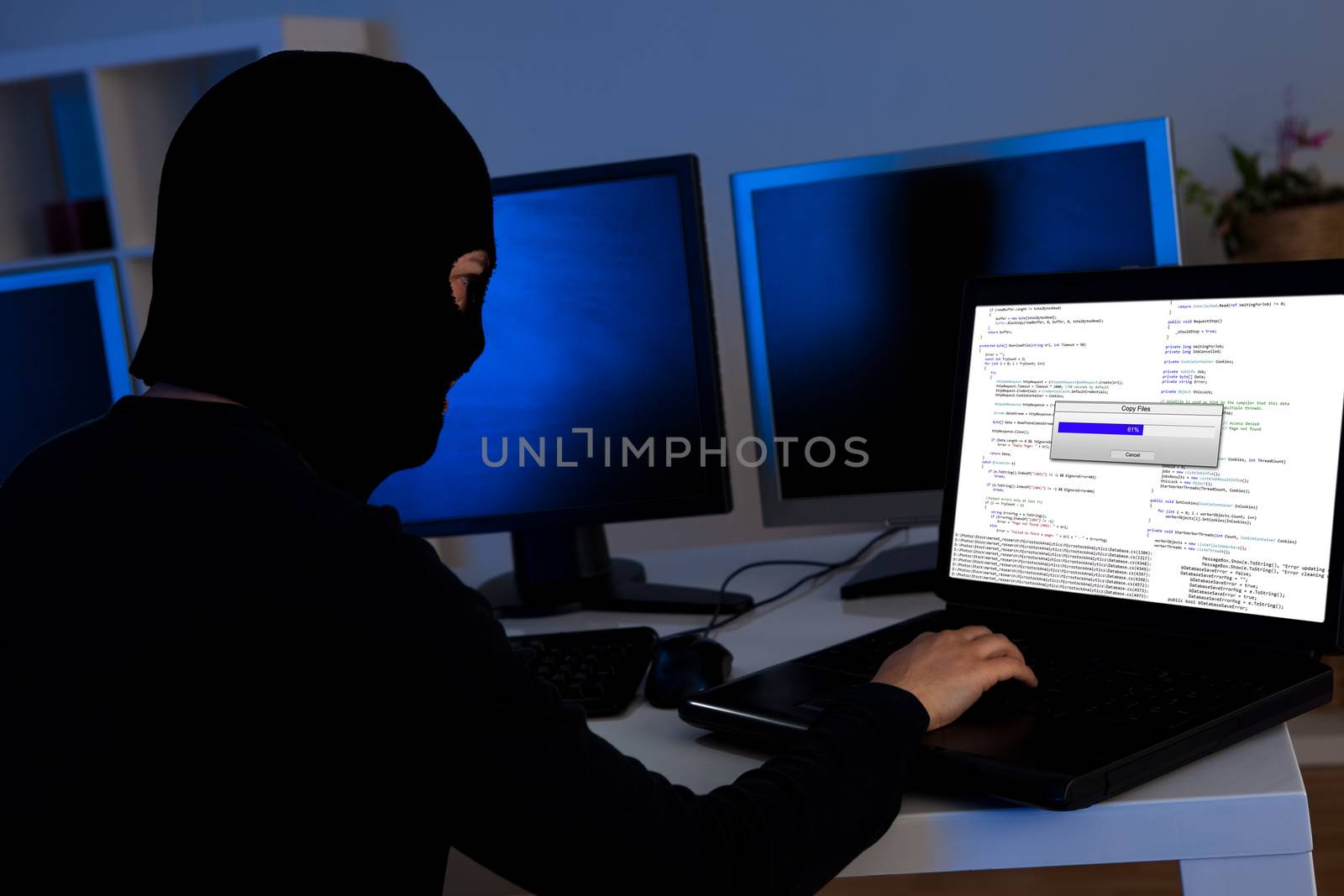 Masked hacker wearing a balaclava sitting at a desk downloading private information off a computer