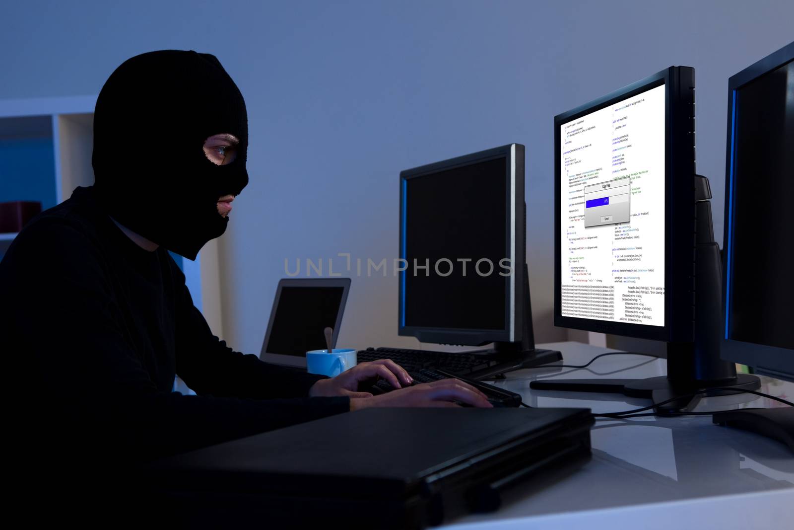 Masked hacker wearing a balaclava sitting at a desk downloading private information off a computer