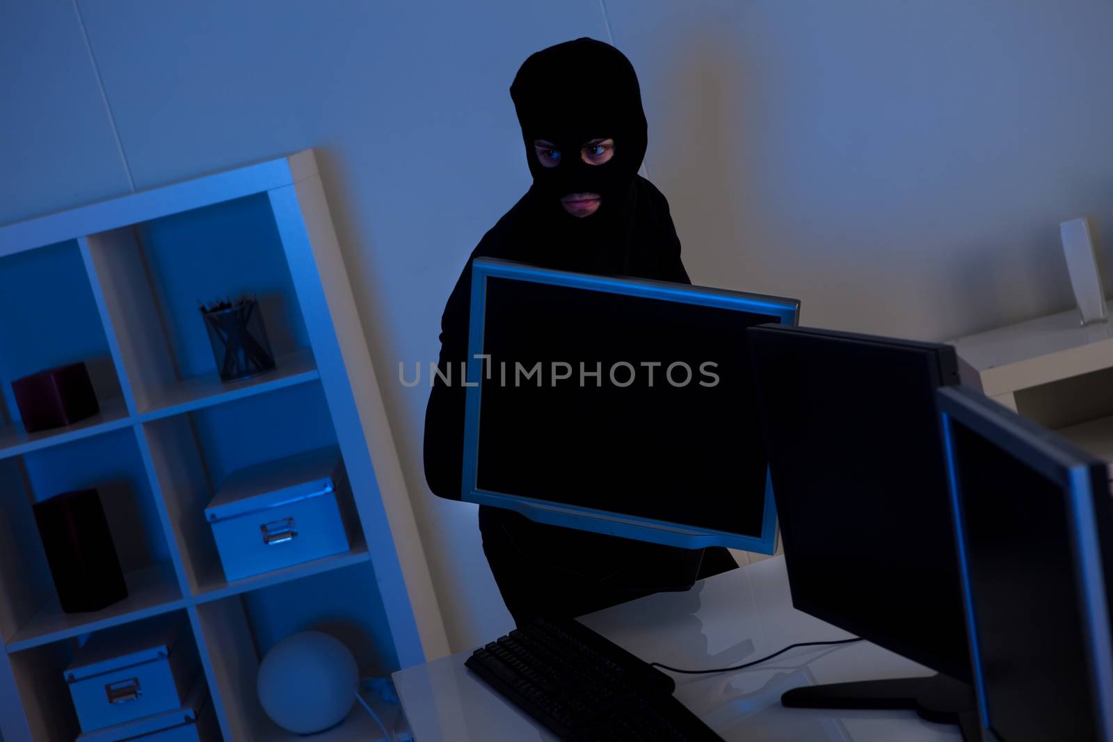 Man dressed in black and wearing a balaclava is stealing a laptop from an office