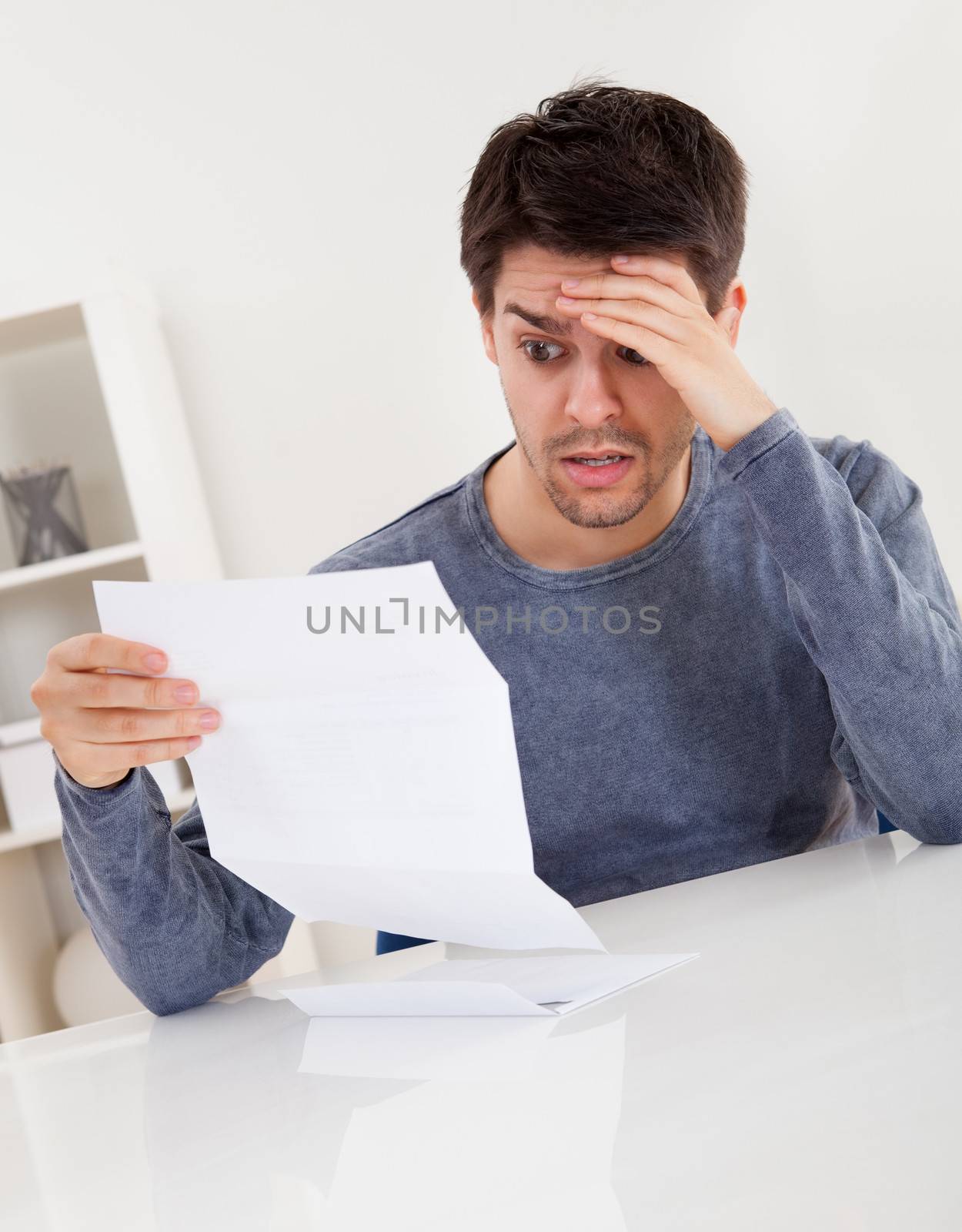 Horrified young man reading a document with an aghast expression and his hand to his forehead as he stares wide eyed at the page of paper