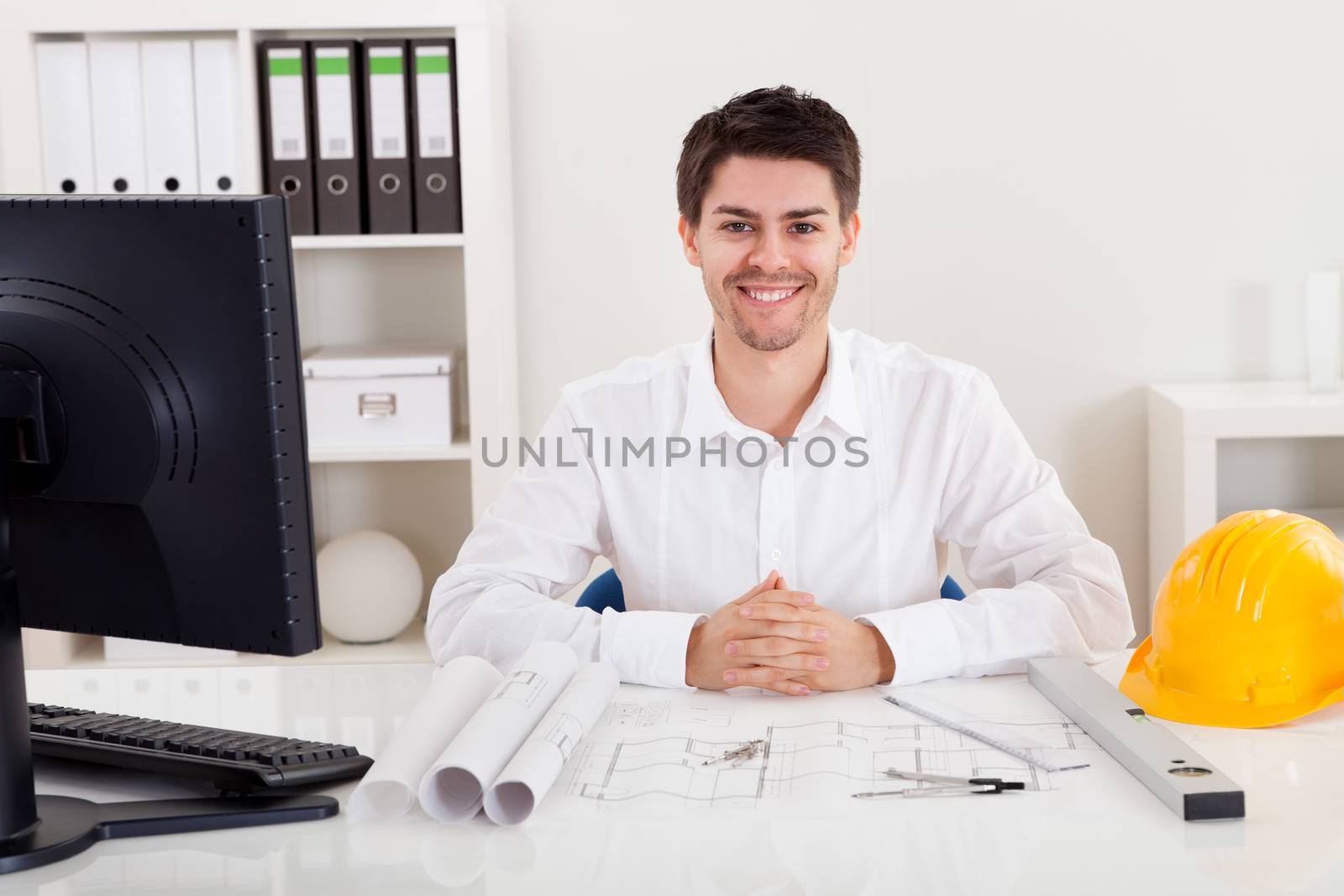 Confident young architect sitting at his desk in his office with a set of rolled blueprints and a hardhat smiling at the camera