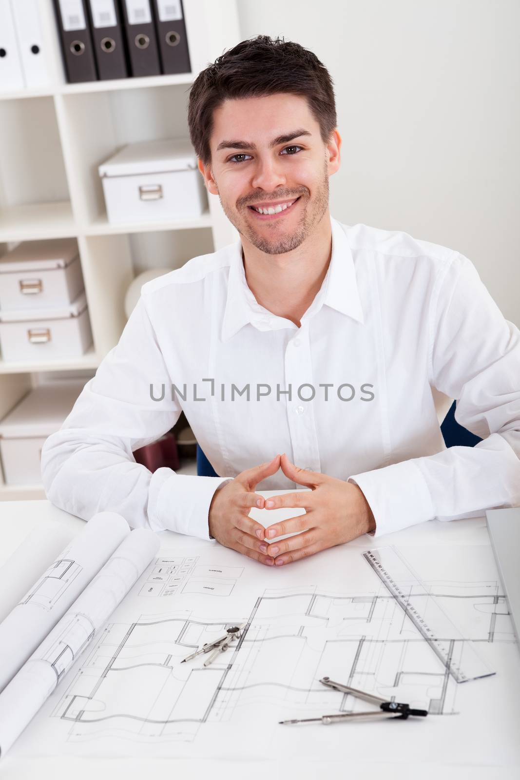 Confident young architect sitting at his desk in his office with a set of rolled blueprints and a hardhat smiling at the camera