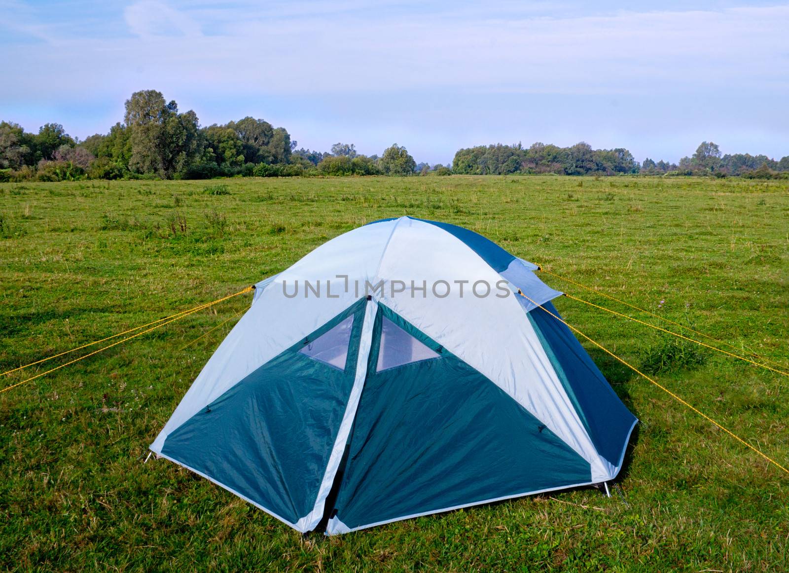 Practical single camping tent of blue-white color on the green meadow by the river.

