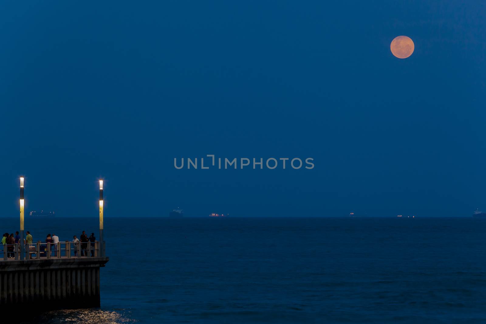 Full moon rising over ocean horizon at twilight with pink red colors with beach pier spectators admiring seasons of nature.