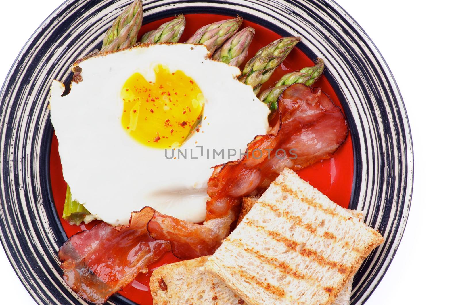 Tasty Breakfast with Fried Egg Sunny Side Up on Delicious Asparagus Sprouts, Roasted Bacon and Toasts on Striped Plate closeup on white background. Top View
