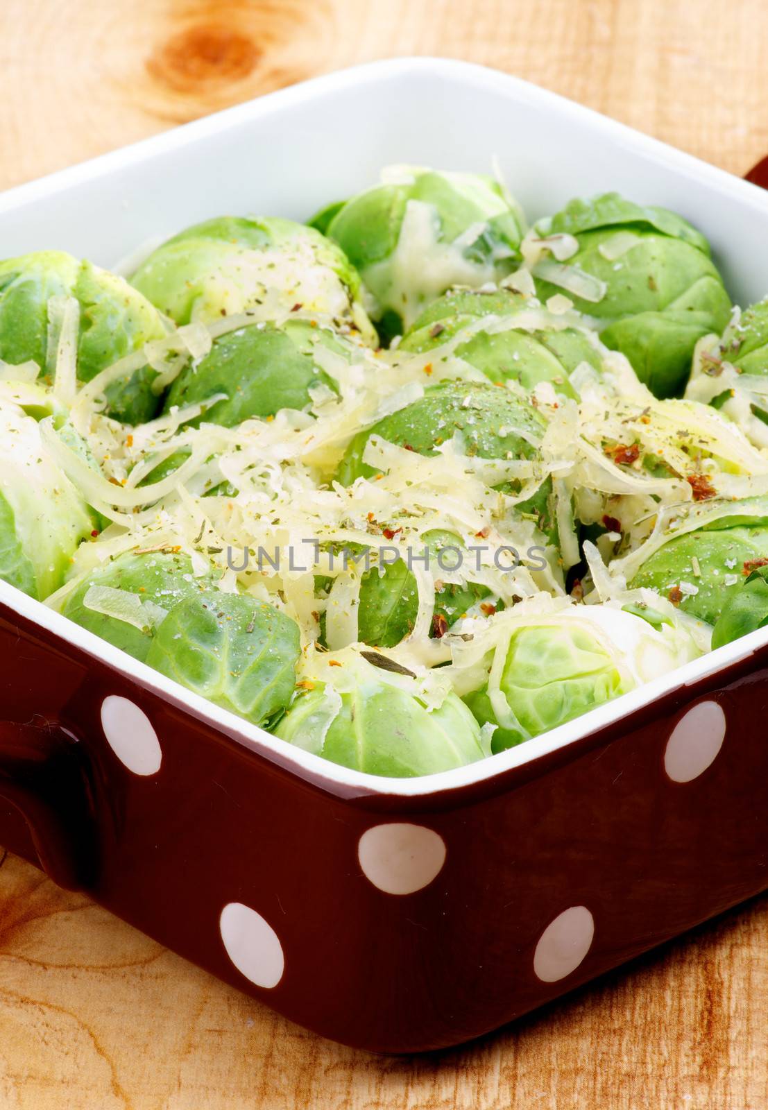 Delicious Homemade Brussels Sprouts Casserole in Brown Polka Dot Bowl with Grated Cheese and Spices closeup on Wooden background