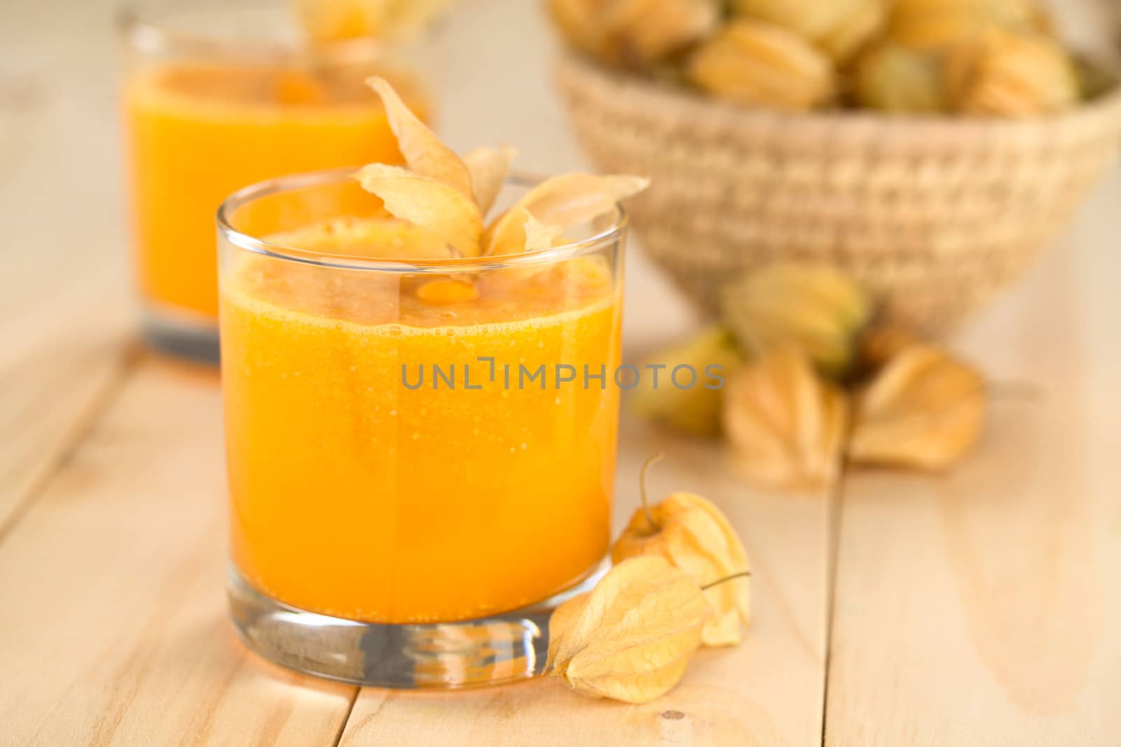 Freshly prepared juice made of physalis (lat. Physalis peruviana) served in glass with physalis fruits on the side and a basket full of physalis in the back (Selective Focus, Focus on the front of the glass rim)
