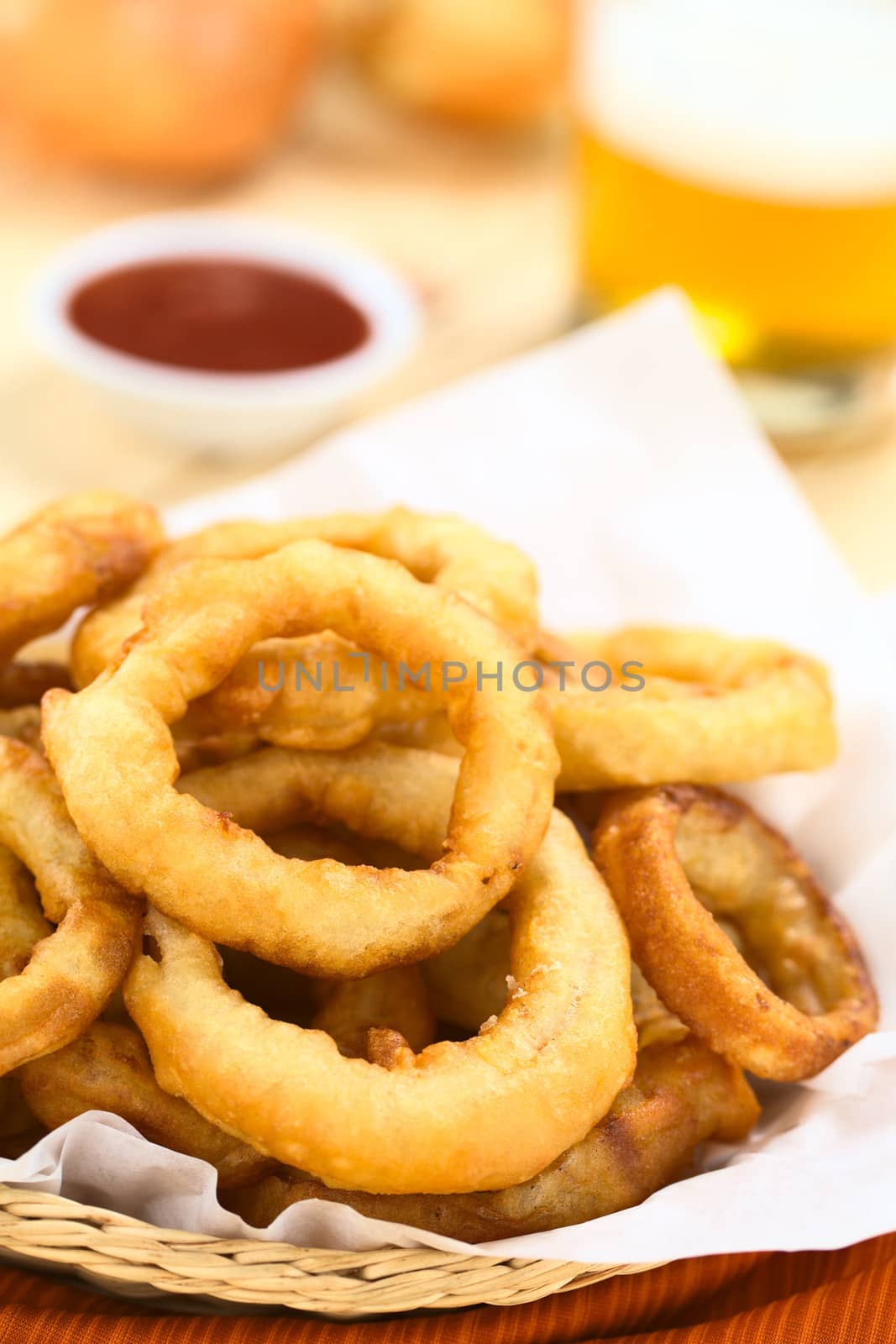 Freshly prepared homemade beer-battered onion rings in a basket with beer and ketchup in the back (Selective Focus, Focus on the front of the onion ring on the top)
