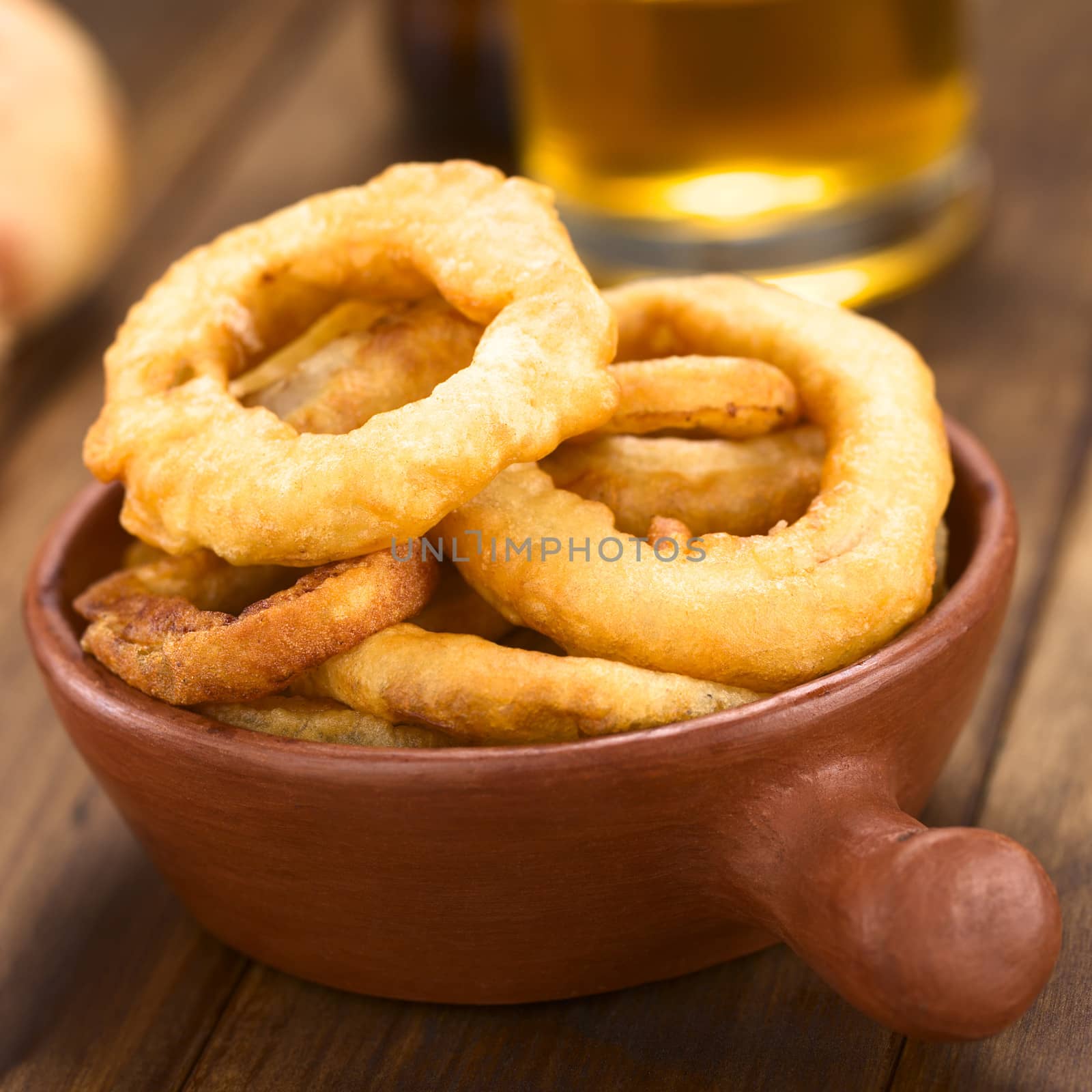 Freshly prepared homemade beer-battered onion rings in a rustic bowl with beer in the back (Selective Focus, Focus on the front of the onion rings on top)